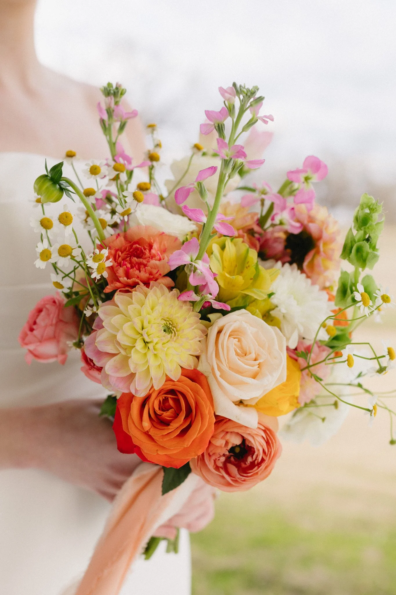 A person holding a colorful bouquet of various flowers including roses, daisies, dahlias, and other blooms with green leaves, wrapped in peach-colored ribbon.