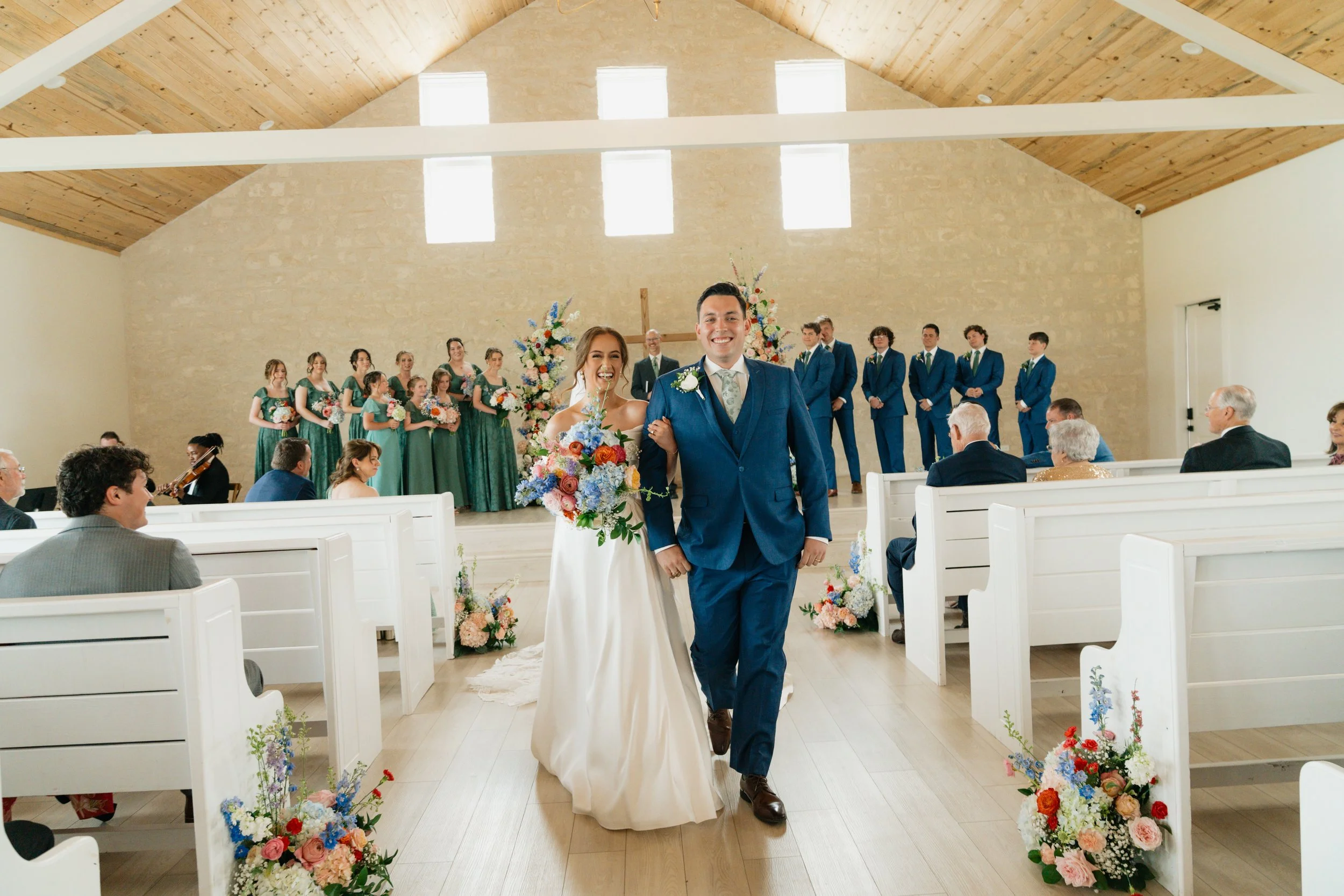 A newlywed couple walking down the aisle in a church during their wedding ceremony, with bridesmaids, groomsmen, and guests present, all smiling and celebrating.