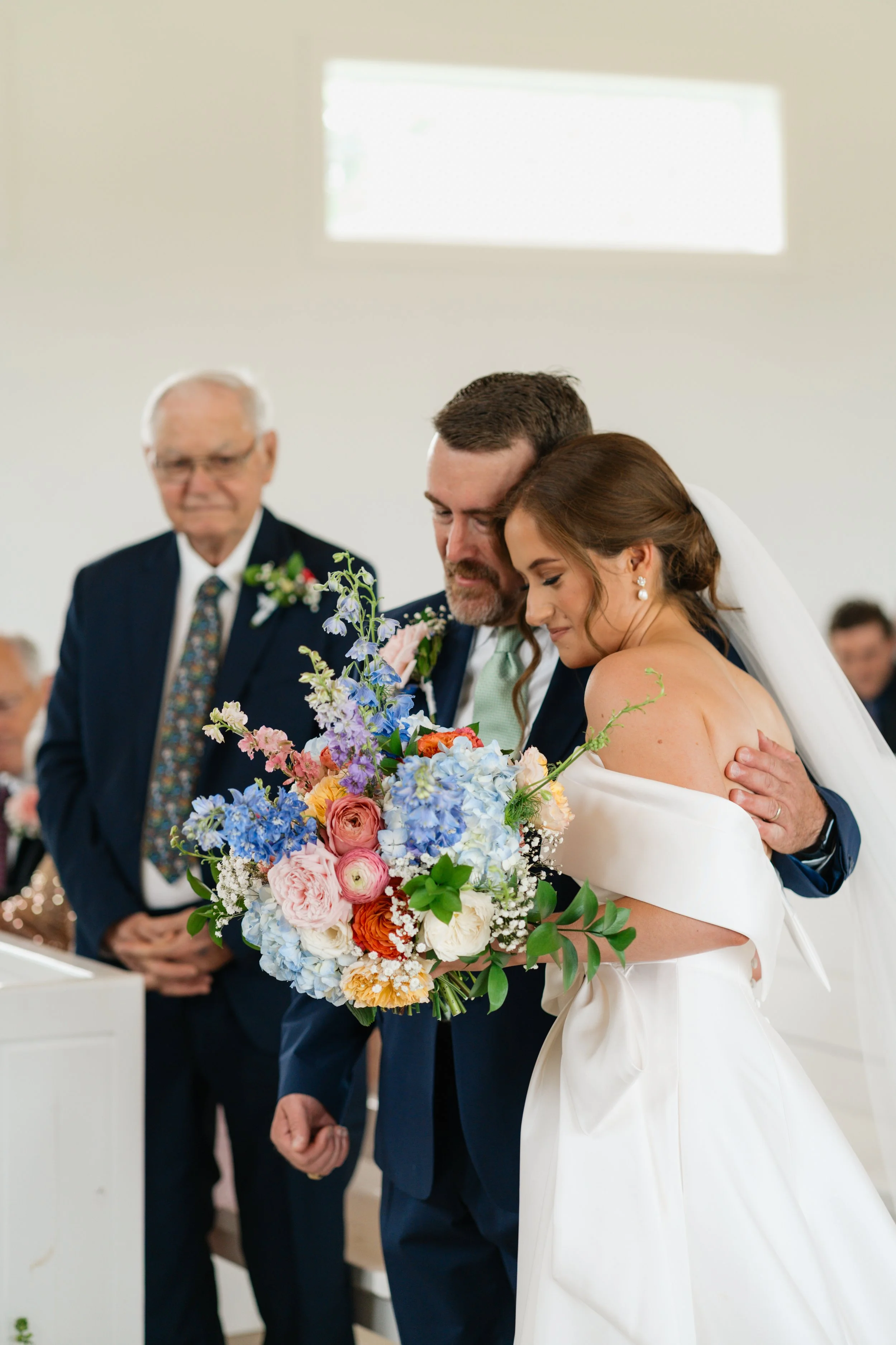 A bride and groom embracing during their wedding ceremony, holding a large colorful bouquet of flowers, with an officiant and guests in the background.