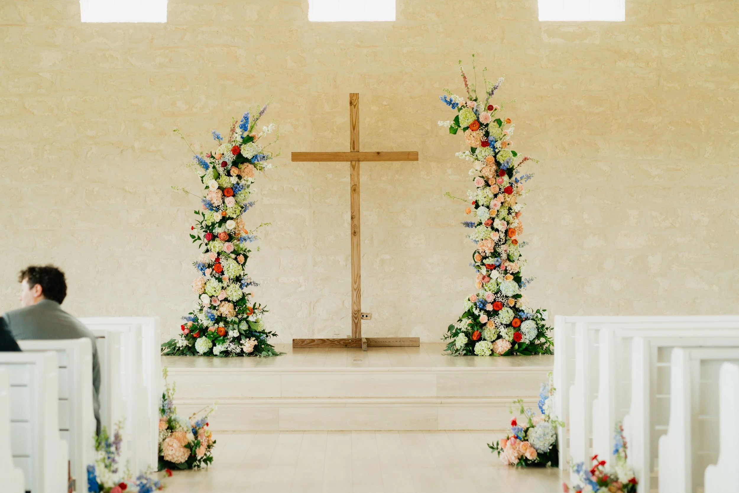 Wedding altar with two large floral arrangements on a wooden stage, cross in the center, and white pews with floral decorations on either side.