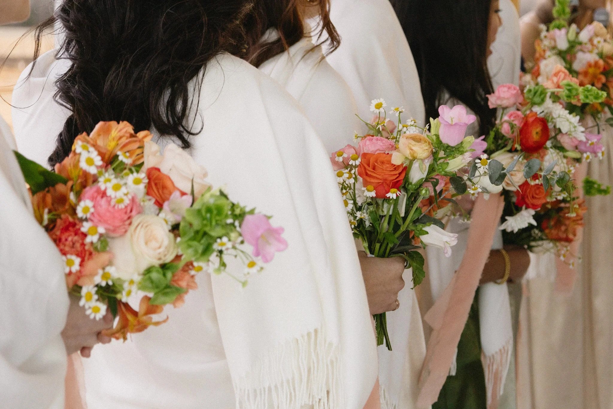 Women dressed in white holding colorful bouquets of flowers, including roses, daisies, and other blooms, during a ceremony.