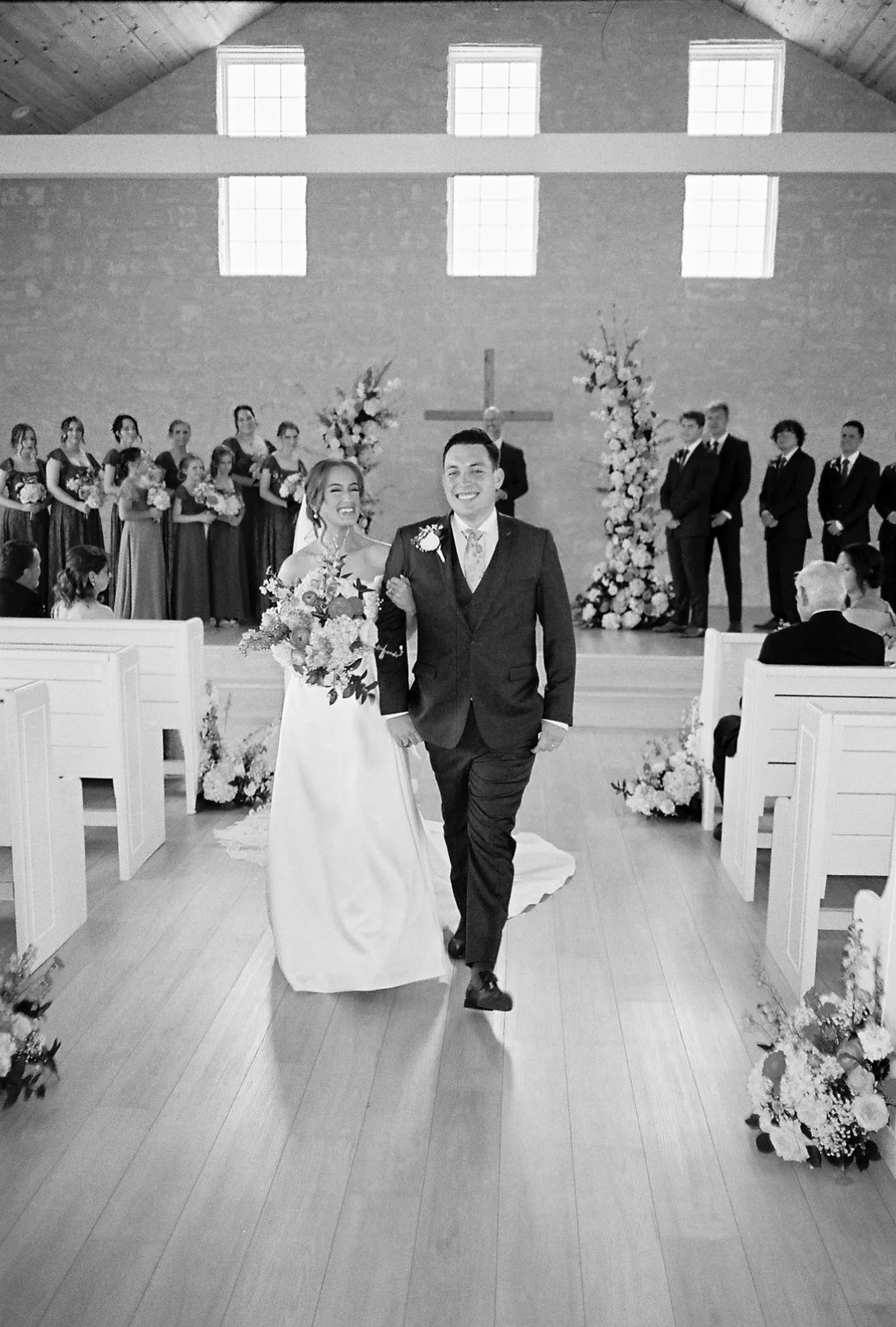 Black and white photo of a bride and groom walking down the aisle in a church, with wedding party and guests in the background.