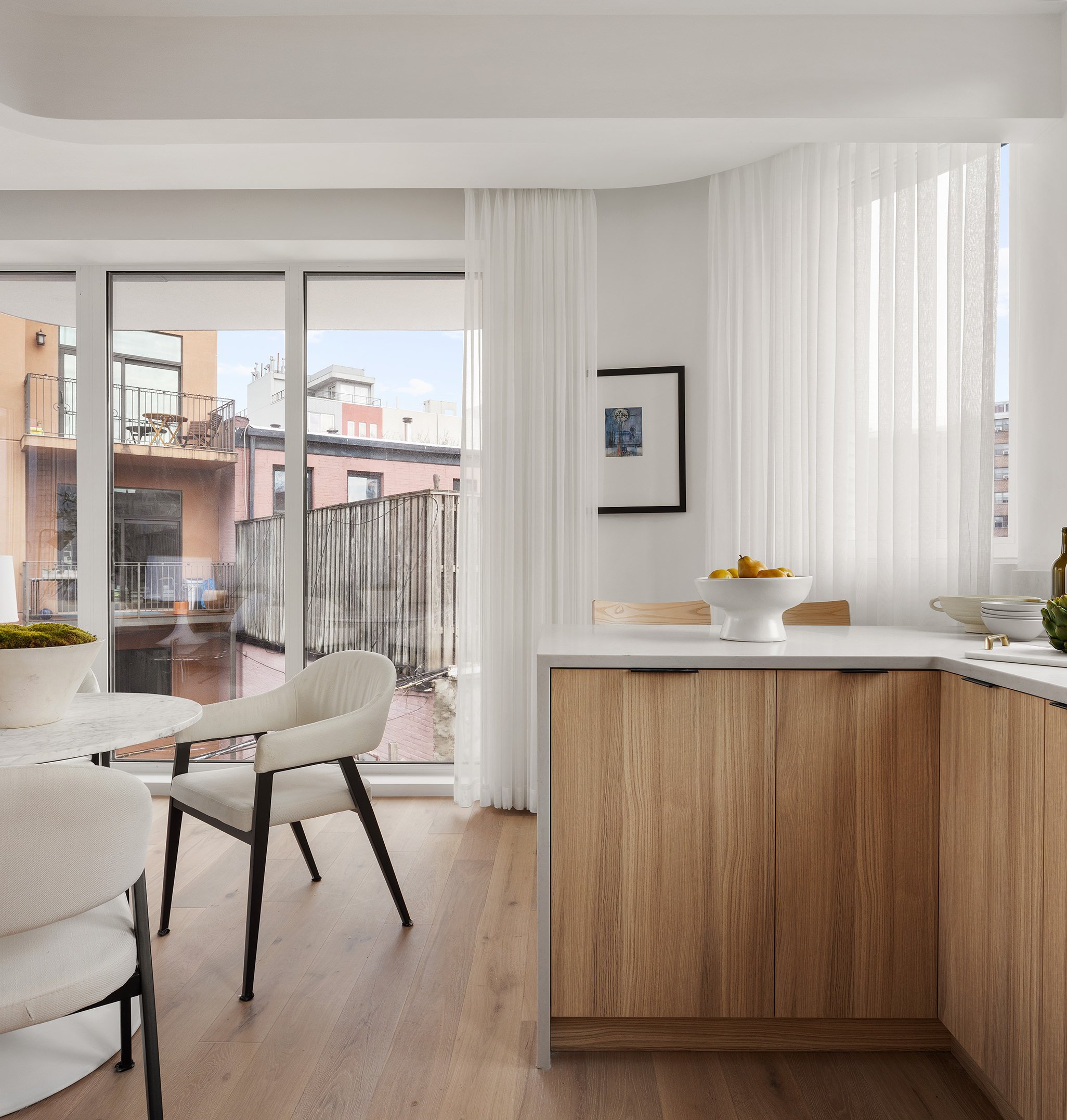 Modern kitchen with wooden cabinets, white countertops, fruit bowl, and sitting area with white chairs and a table, overlooking an urban balcony with buildings.