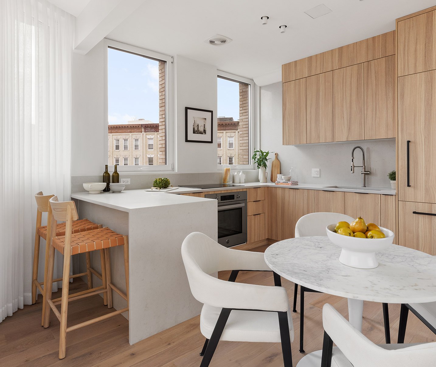 Modern kitchen with wooden cabinets, white countertops, and a round marble dining table with white chairs. There are two large windows, a wall art piece, and a bowl of green apples on the table.