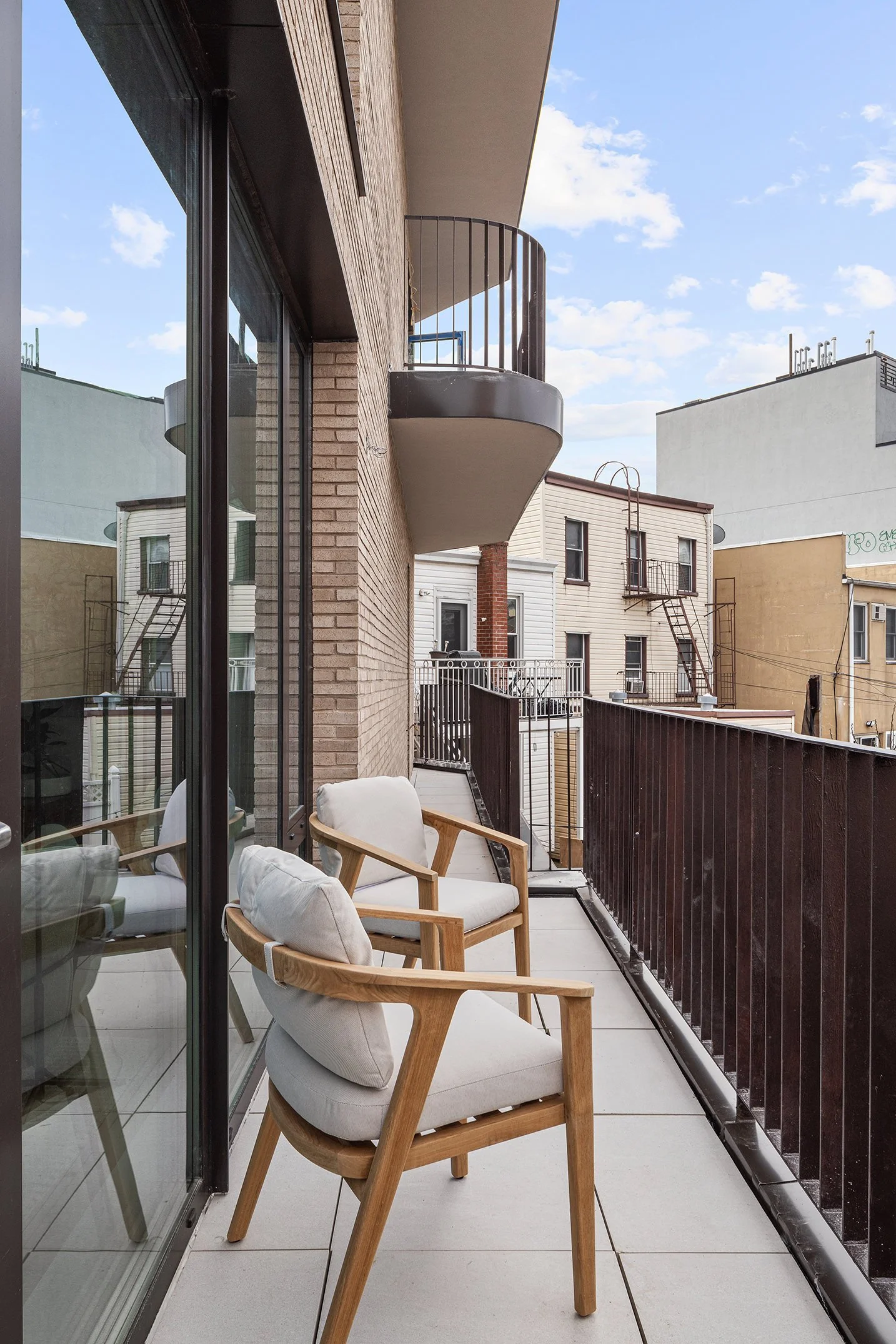 Balcony with two white cushioned chairs and a glass sliding door, overlooking neighboring buildings and a blue sky with some clouds.