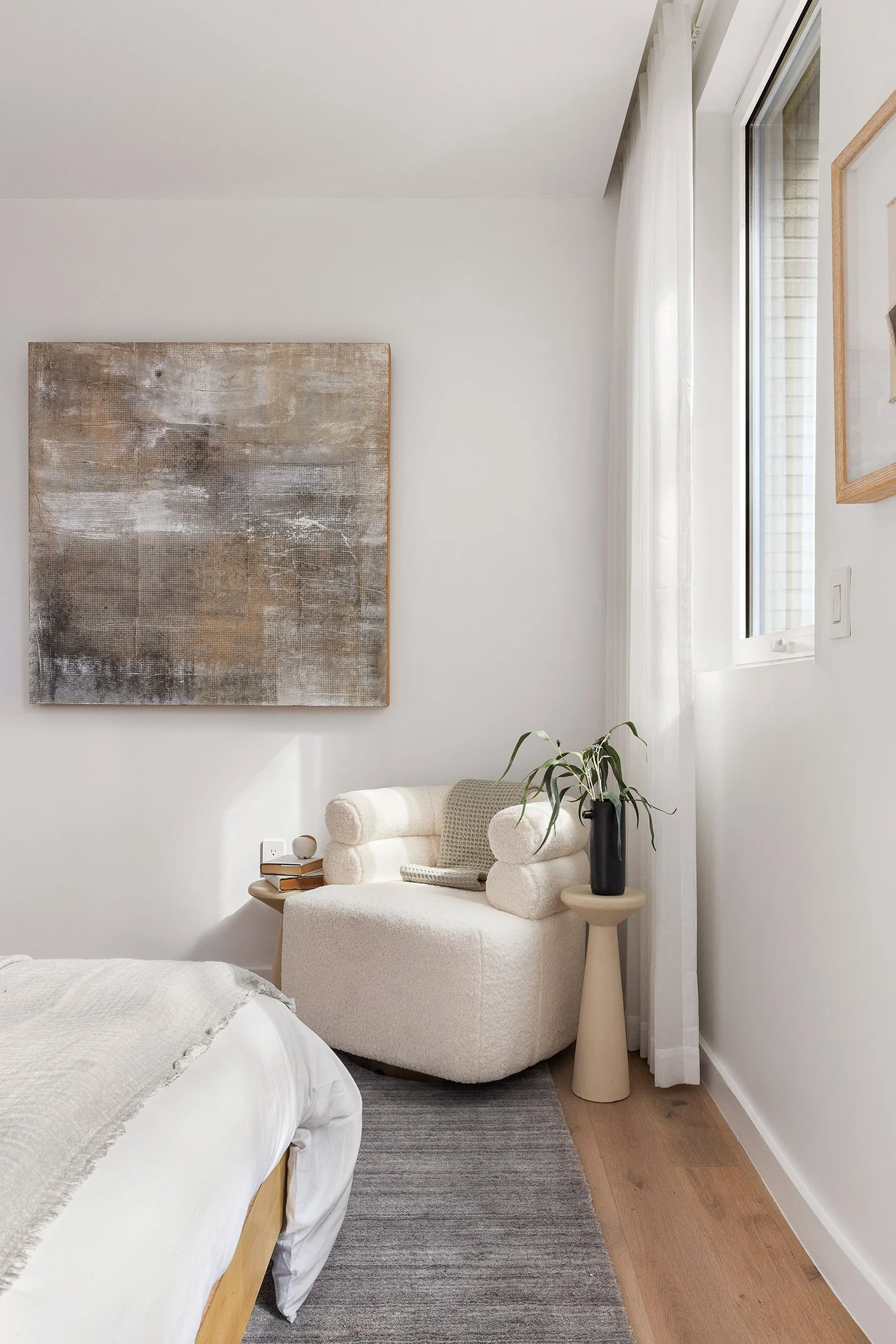 A cozy bedroom corner with a white armchair, stack of books, a black vase with a plant, and wall art next to a window with white curtains.