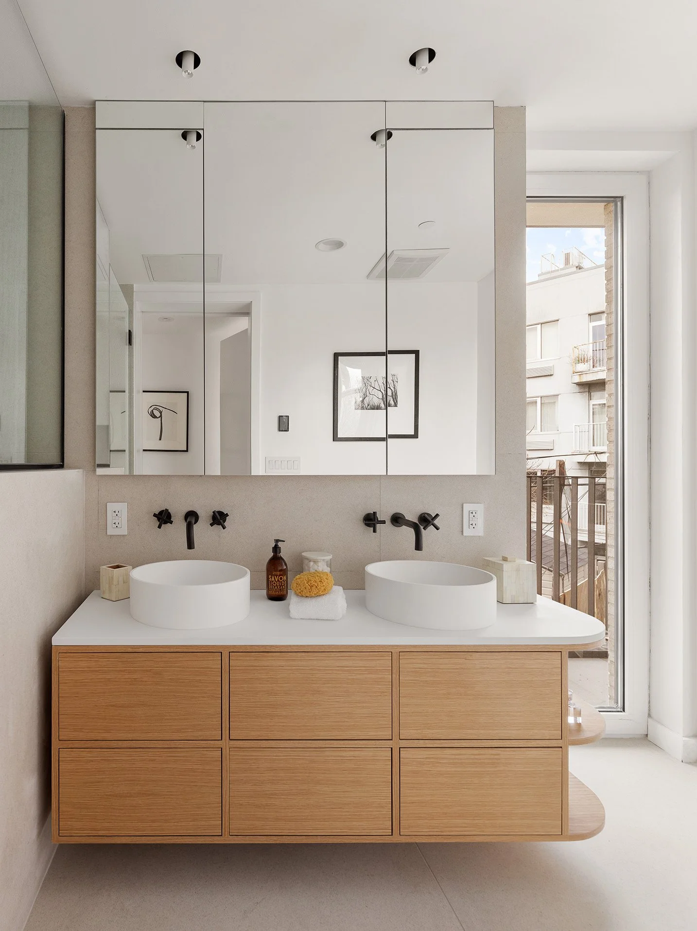 A modern bathroom with a double vanity featuring two vessel sinks, black wall-mounted faucets, and a large mirror. Decor includes a soap dispenser, a sponge, a candle, and tissue boxes. A balcony with an outdoor view is visible through a glass door o