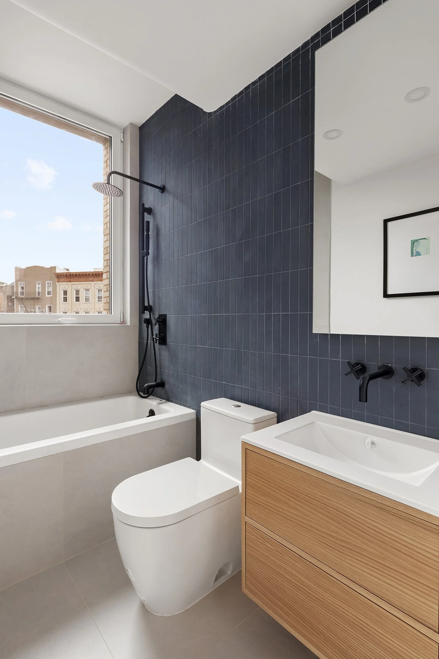 Modern bathroom with white bathtub, wall-mounted black shower, and toilet. A wooden vanity with a white sink is next to a large mirror, with a window providing natural light. The bathroom features dark blue tile walls and light-colored floor tiles.