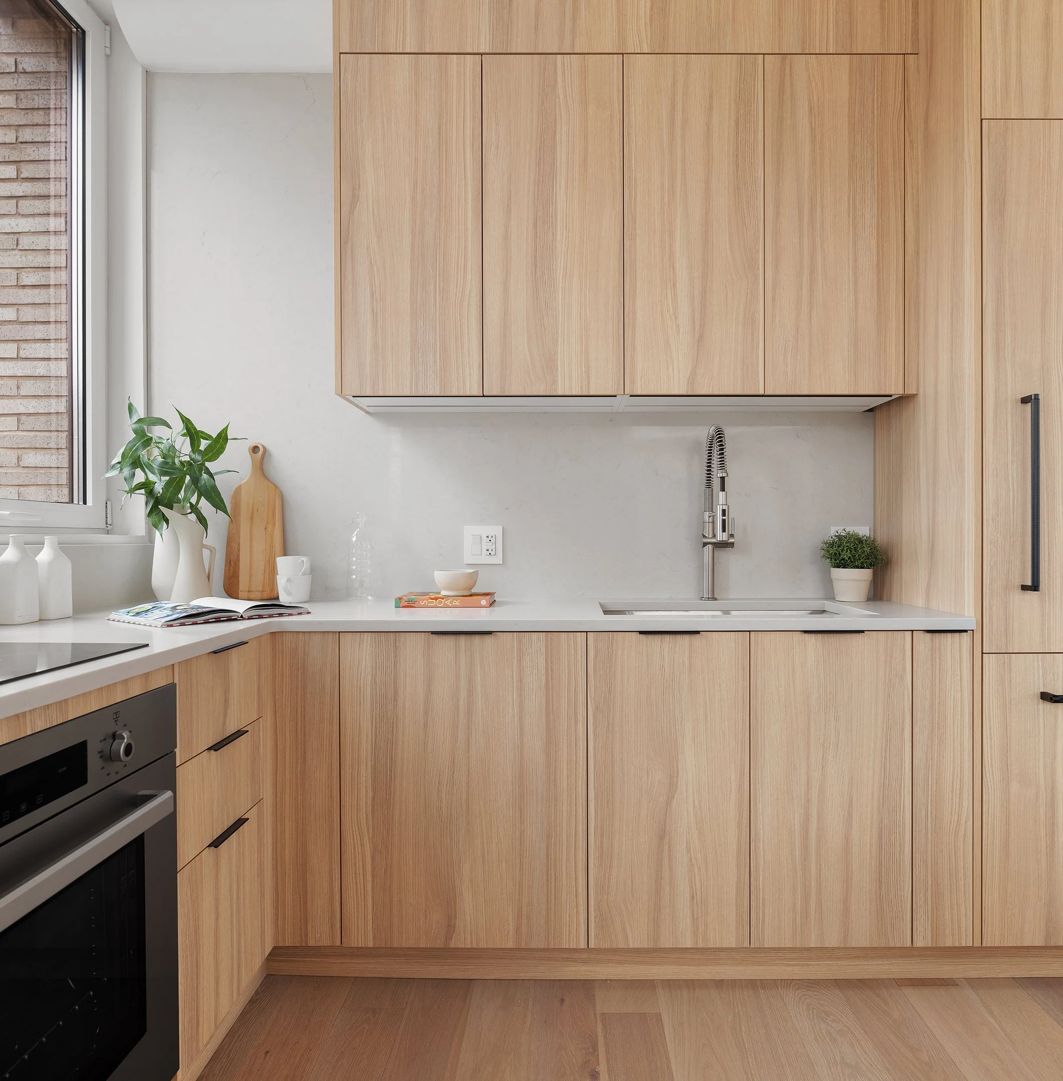 Modern kitchen with light wood cabinets, white countertop, stainless steel sink with a high-arc faucet, and minimal decor including a potted plant, books, and decorative vases.