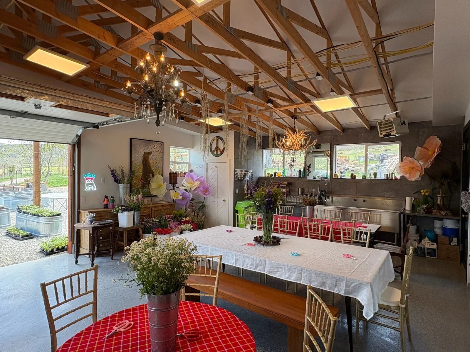 Interior of a decorated event space with a large table covered with a white tablecloth, surrounded by gold chairs. The room has an open garage-style door, revealing outdoor seating and greenery. The ceiling features exposed wooden beams and chandelie