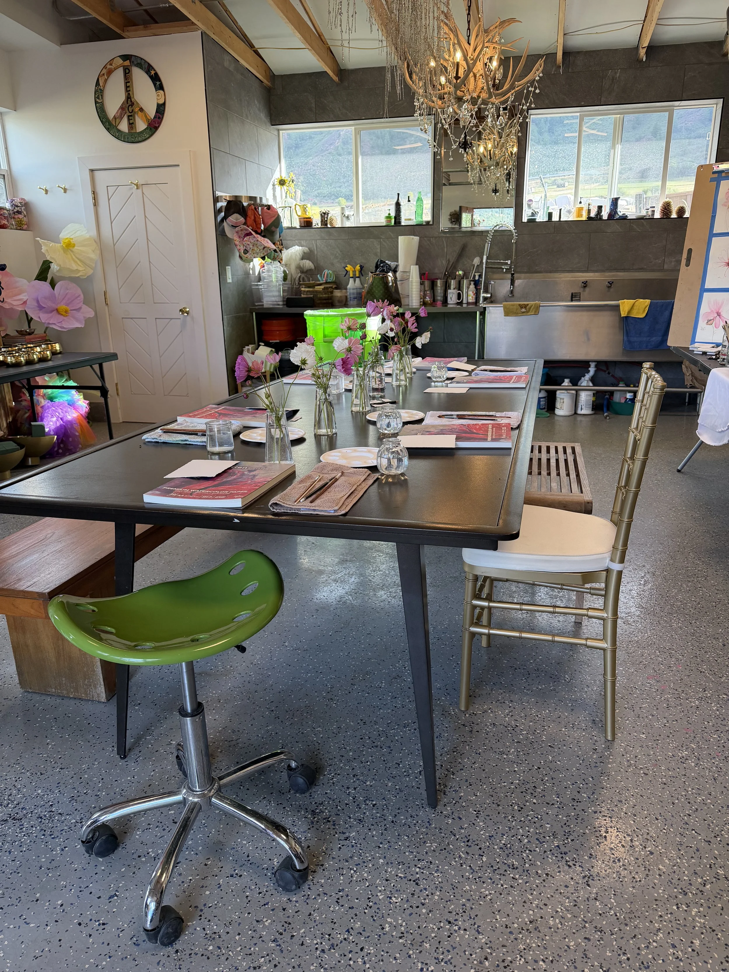 Indoor meeting or workshop space with a long table set with notebooks, plates, and glasses. Decorative flowers in vases, colorful chairs including a green one on wheels and a gold one, and a chandelier hanging from the ceiling. Large windows show an 