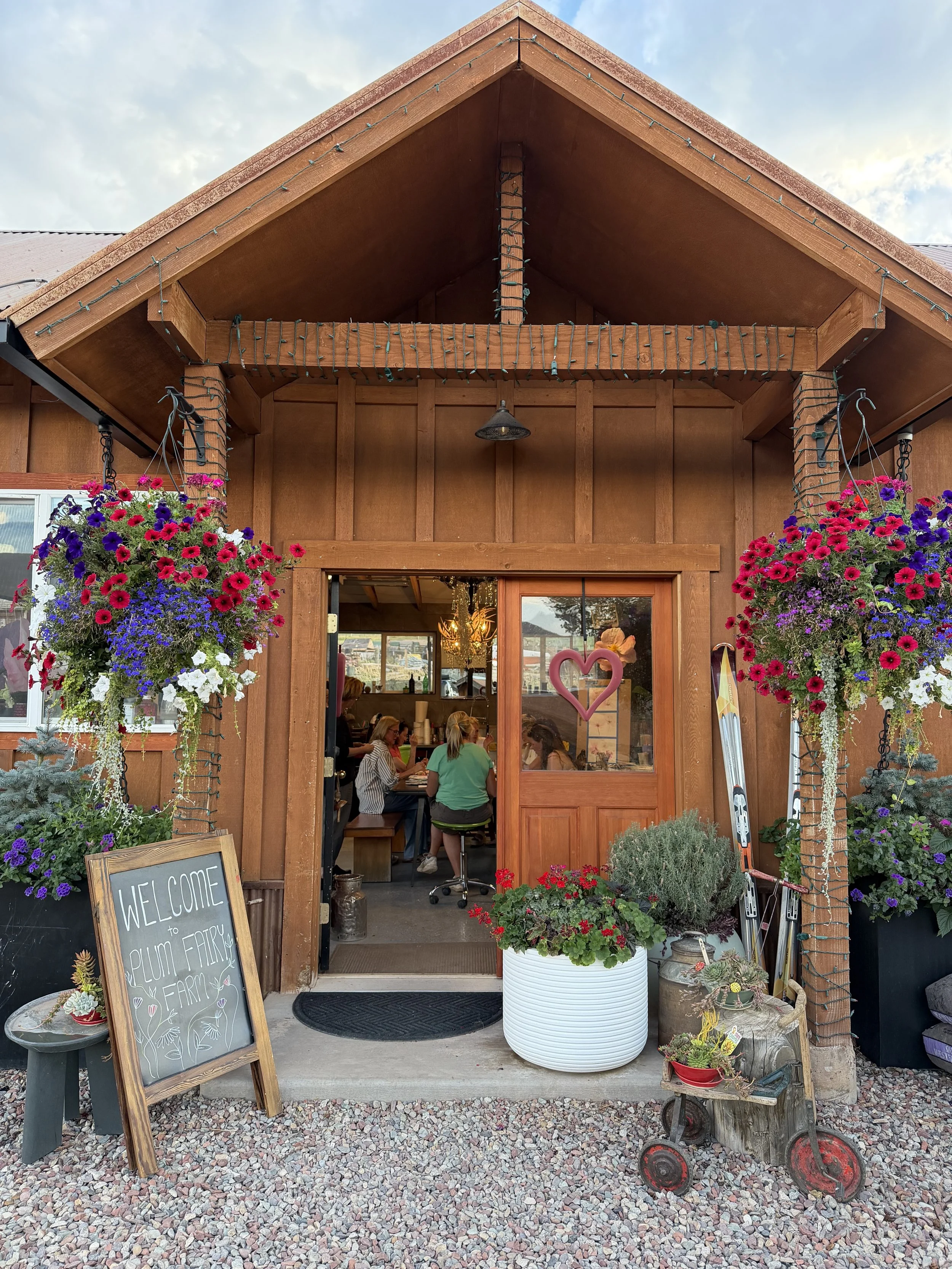 The entrance of a farm shop with hanging flower baskets on each side and plants in pots outside. A chalkboard sign reads welcoming visitors to Plum Fairy Farm. Inside, people are sitting at tables. A heart-shaped decoration hangs on the door window. The farm shop has wooden walls and decorated with outdoor plants and rustic items.