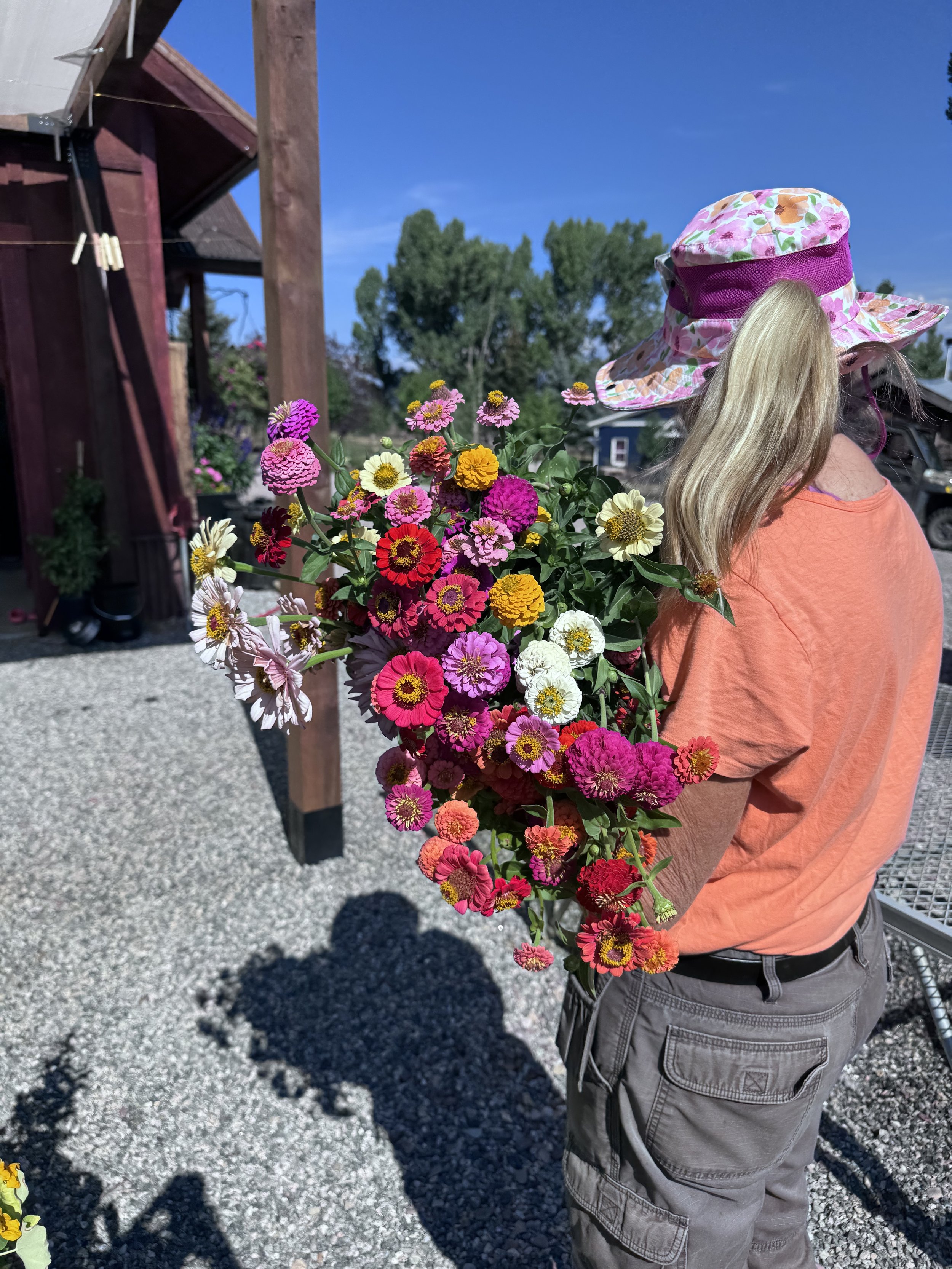 A child wearing a pink floral hat and orange shirt, holding a large bouquet of colorful flowers, standing outdoors on gravel with green trees and a blue sky in the background.