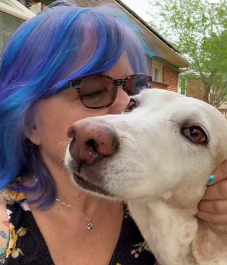 Woman with blue hair, glasses, and a necklace kissing a white dog outside.