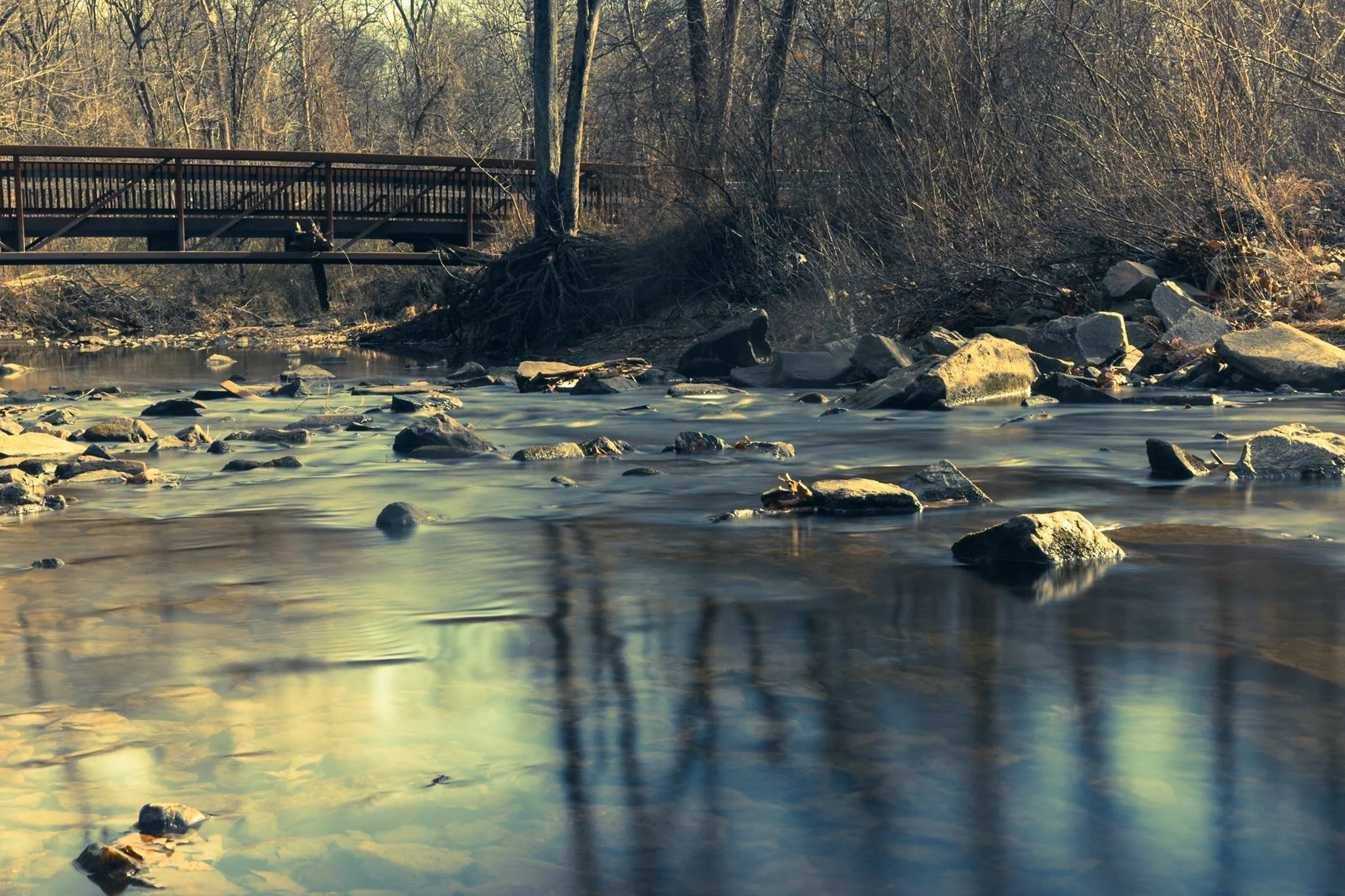 Timed exposure photography of a creek with a walking bridge tinted in blue/ yellow