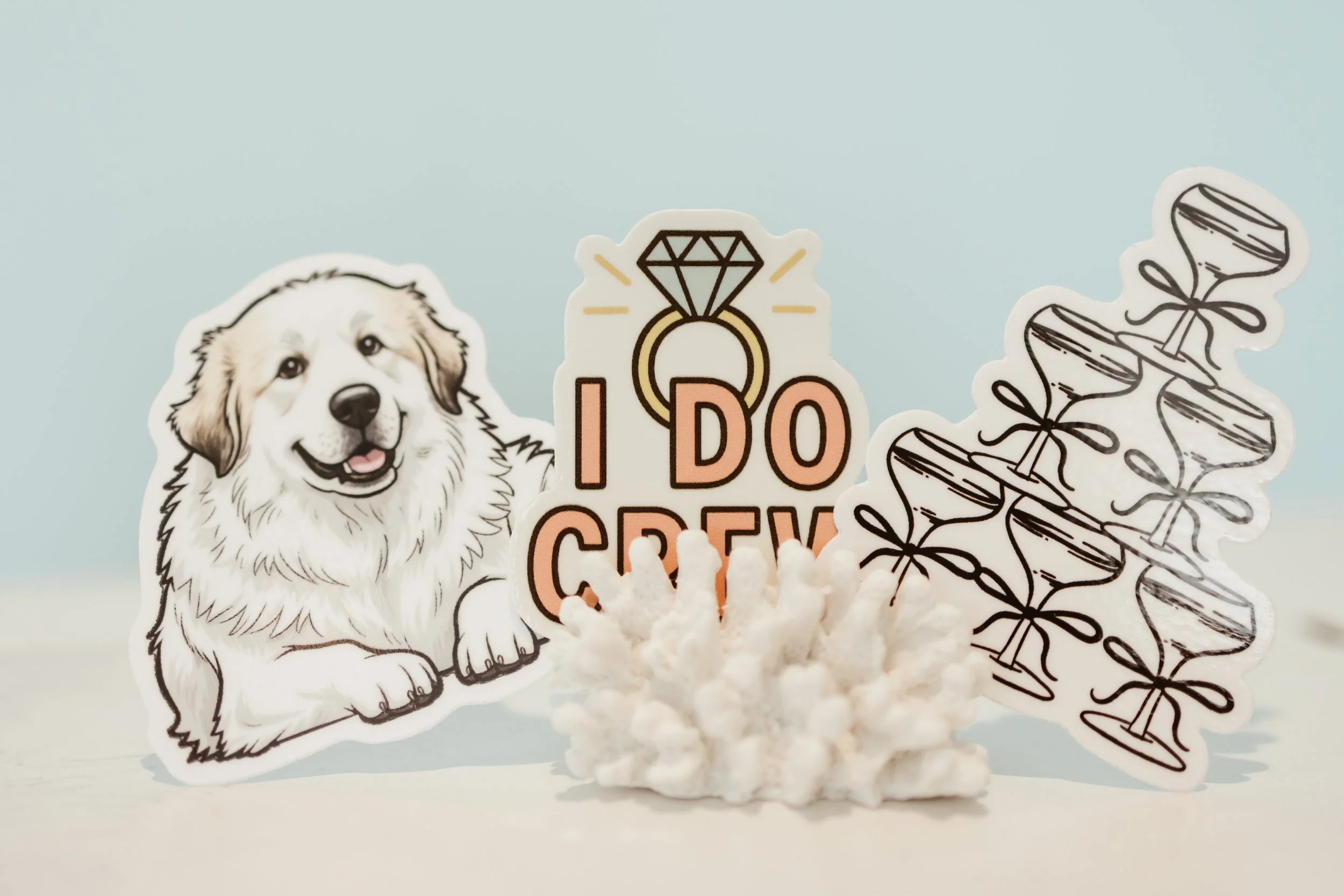 Dog-themed stickers with a happy dog, the phrase 'I do crew' with a diamond ring, and a tower of martini glasses tied with bows, arranged with a white coral piece on a light blue background. - Image by BG Studios 