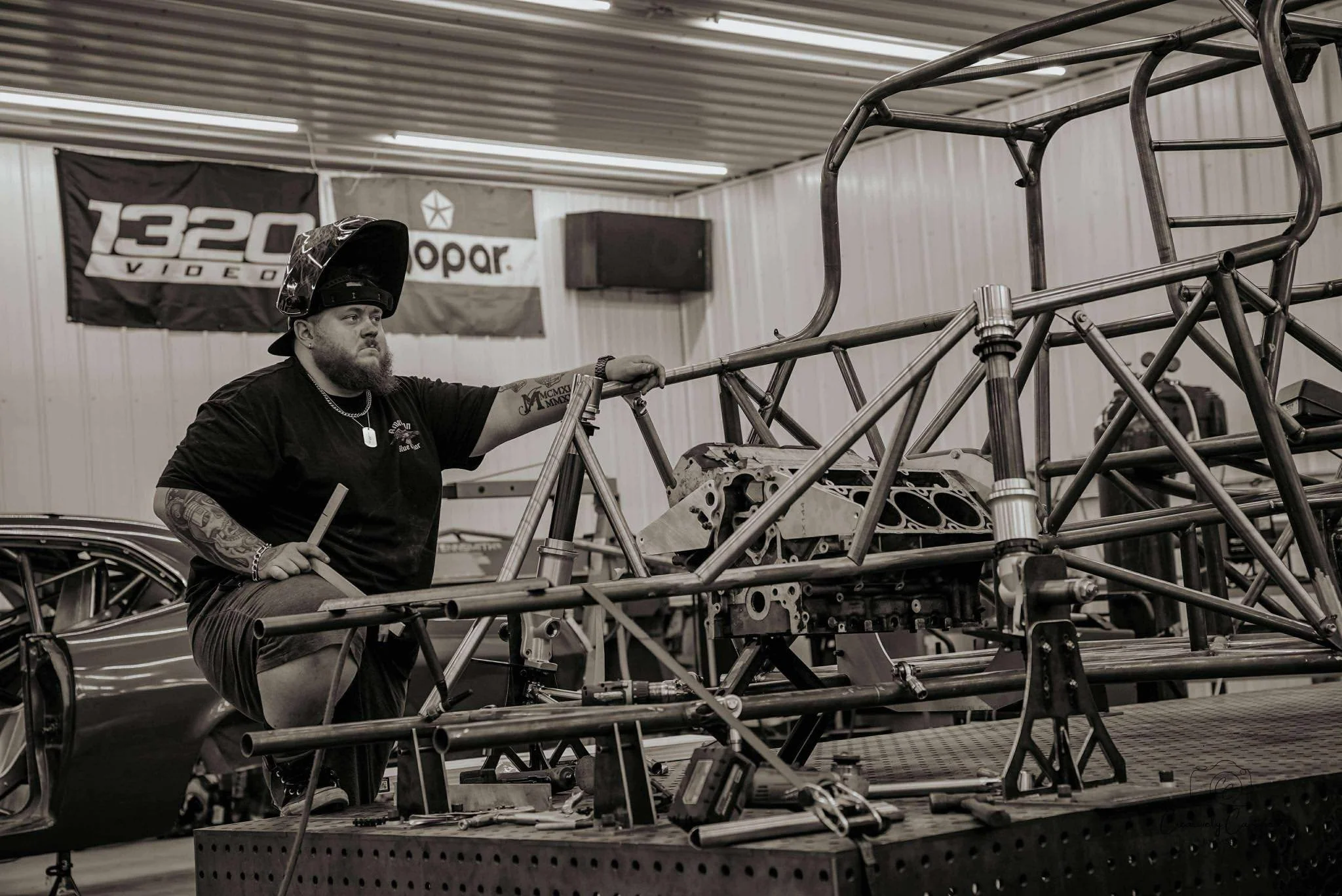 A man wearing a welding helmet, black t-shirt, and shorts working on a metal car chassis in a garage with banners on the wall.