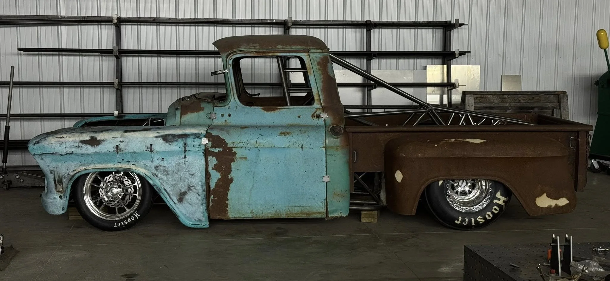 A vintage pickup truck body with a rusted and weathered blue and brown paint job, parked indoors on a concrete floor with metal shelving in the background.