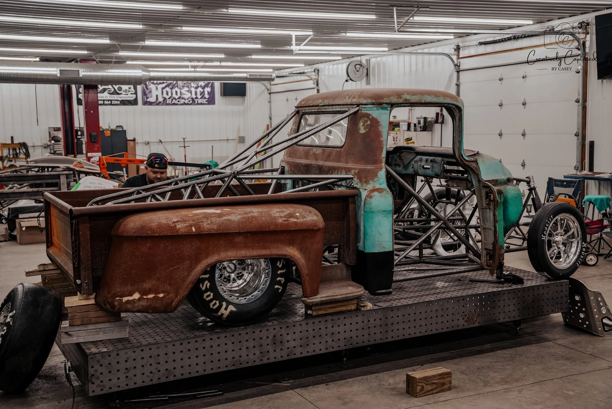 An old, rusty vintage pickup truck frame with a teal and Rusted body, missing parts, on a metal platform in a garage or workshop.