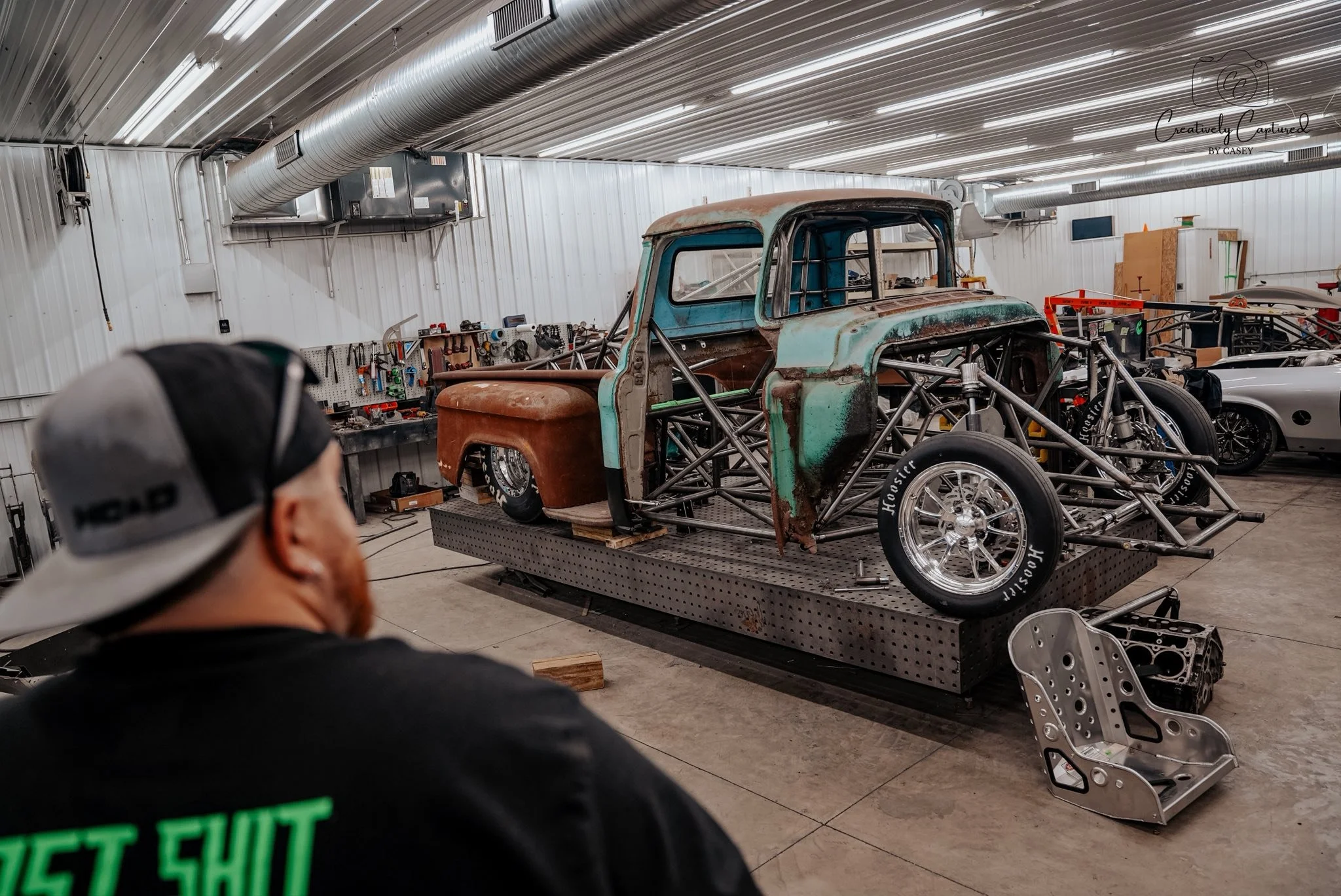An unfinished vintage race car with exposed metal frame and rusted body in a workshop, with a man in a cap and black shirt looking at it.
