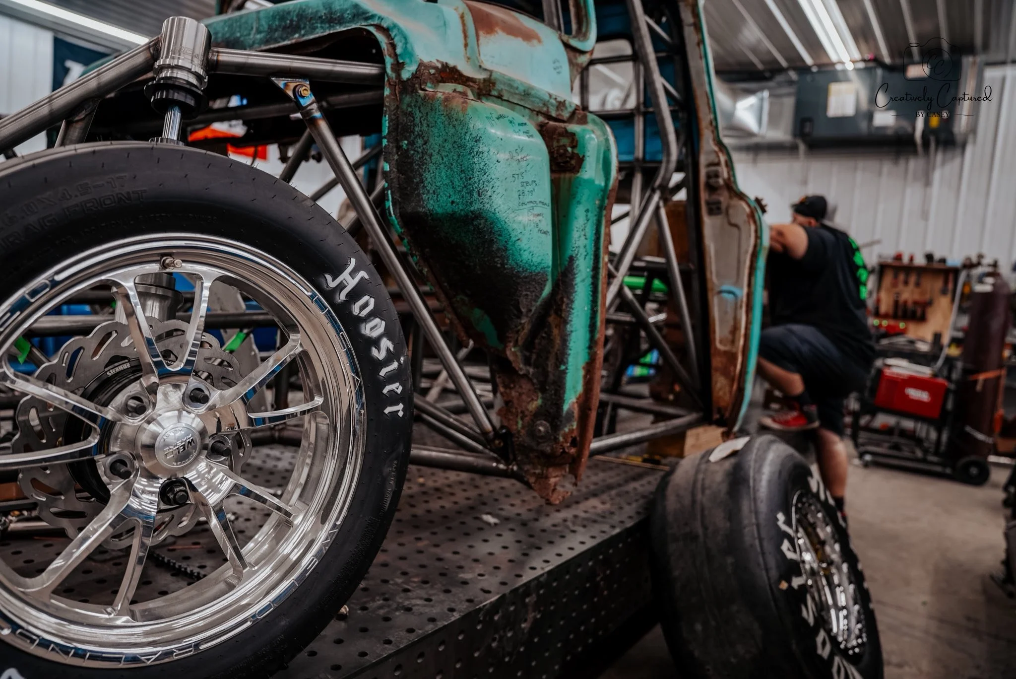 Close-up of a race car's front wheel with a Hoosier tire and a polished rim, with the car's body showing rust and green paint, inside a garage with a person working in the background.