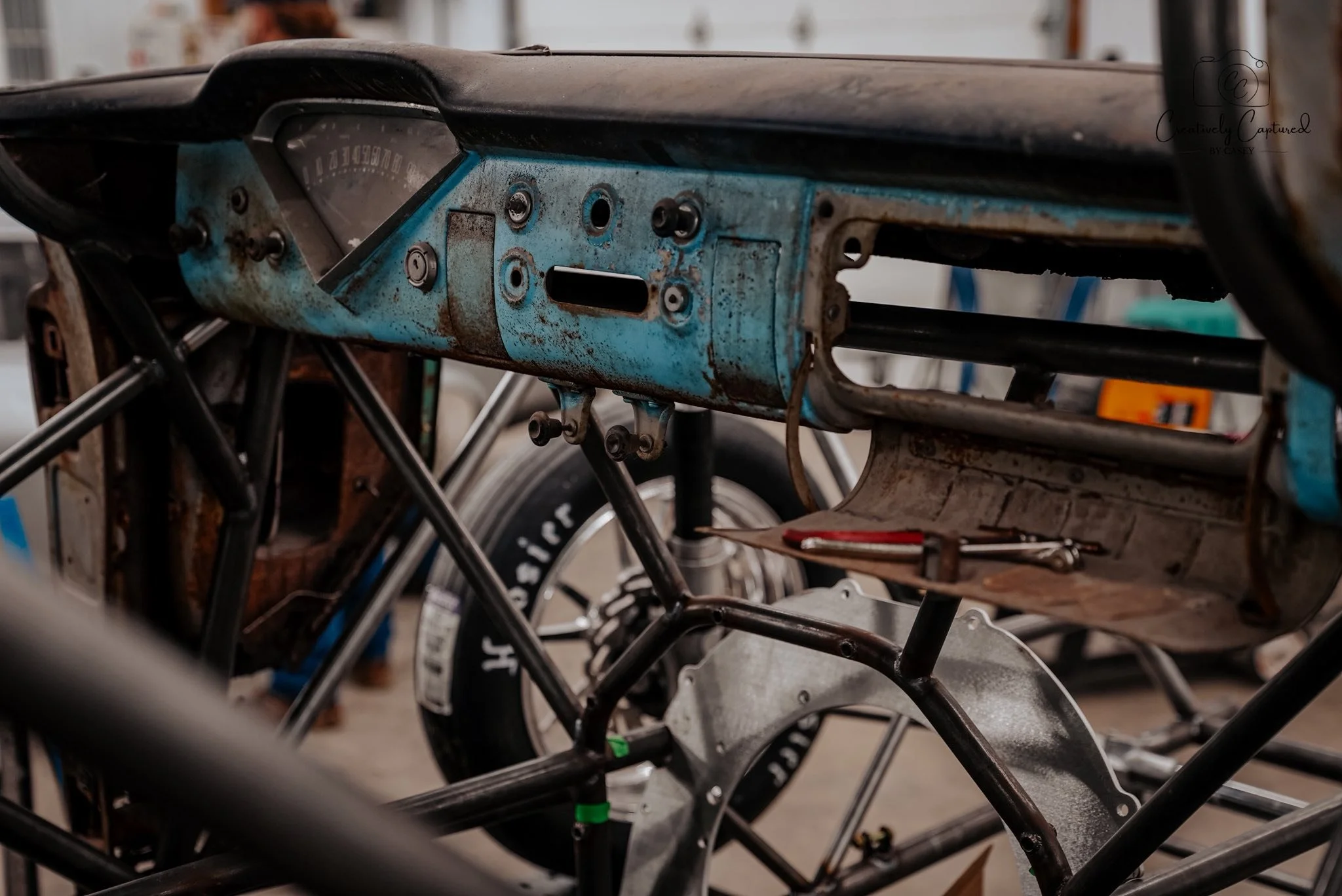 A metal dashboard and frame of a vintage or partially disassembled vehicle, with a rusted blue panel, exposed bolts, and wheel components visible.