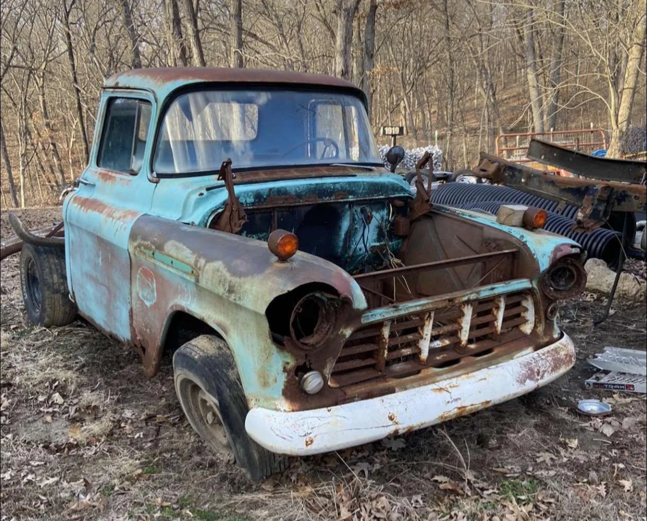 An old, rusted pickup truck with no front grille and missing headlights, parked in a wooded area surrounded by fallen leaves and debris.