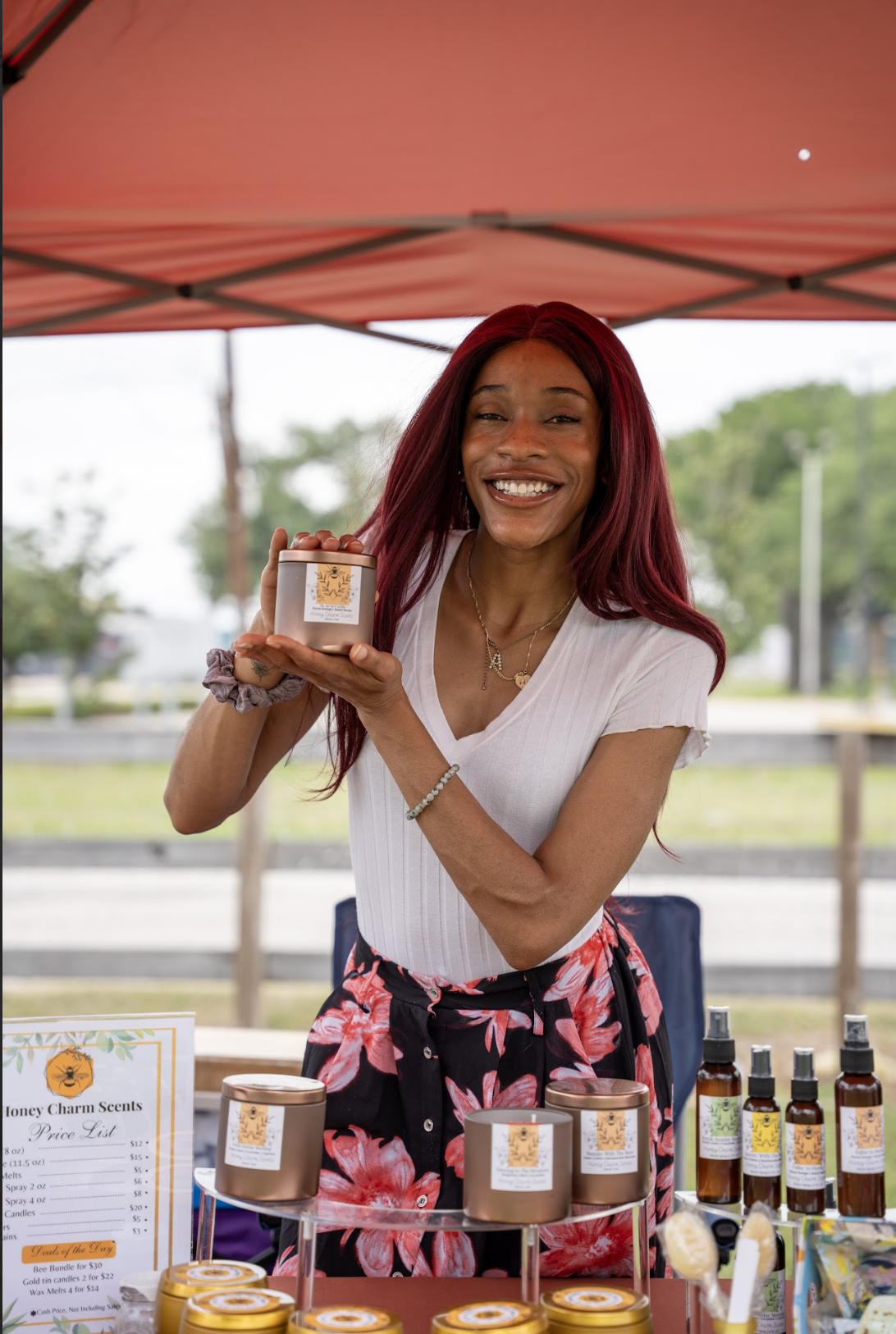 Smiling woman at an outdoor market holding a scented candle in a jar, with various candles and spray bottles on the table in front of her.