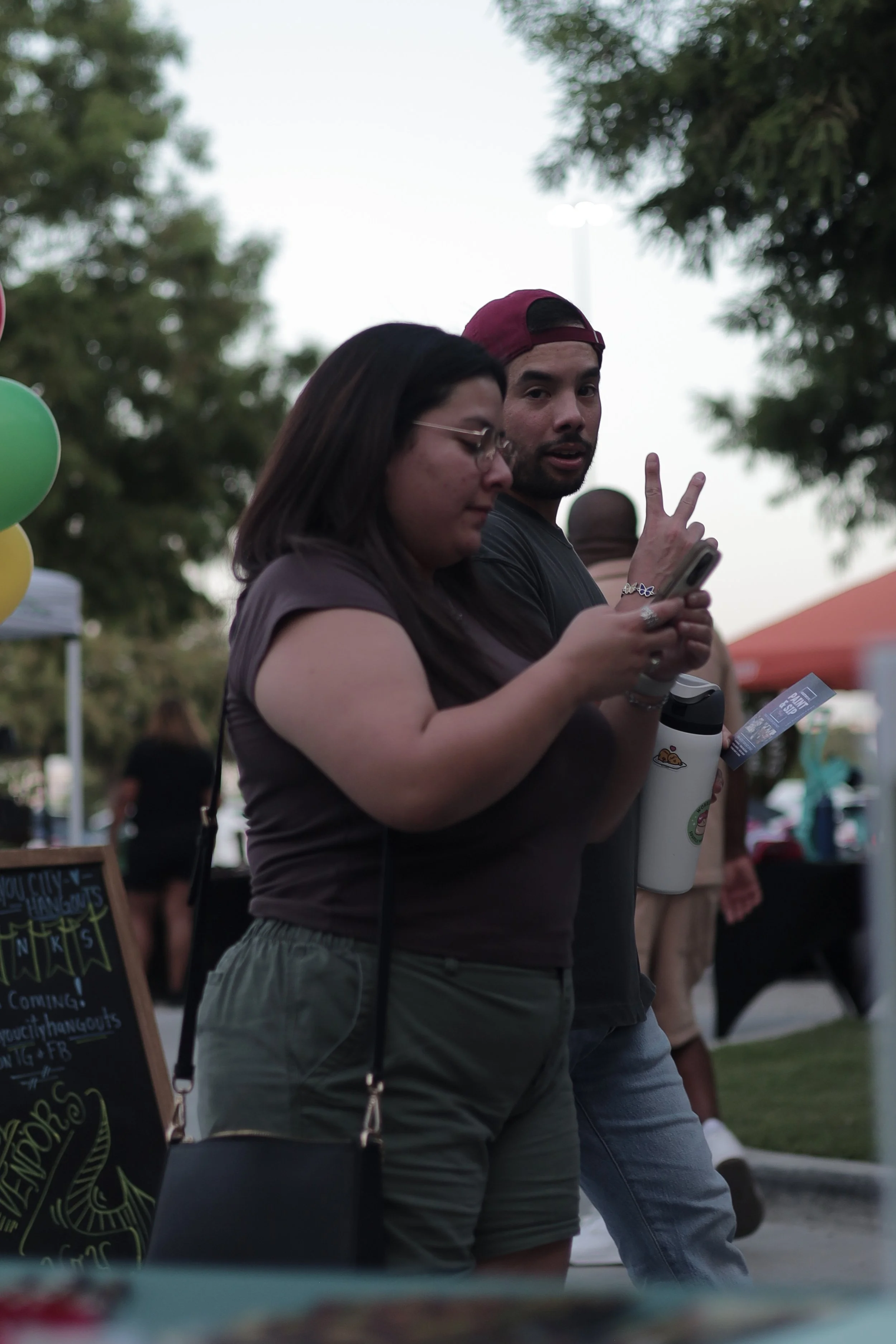 A woman with dark hair and glasses wearing a brown t-shirt and green shorts is looking at her phone, standing next to a man with a beard wearing a backwards cap, black t-shirt, and light blue jeans. The man is making a peace sign with his hand and looking at the camera, holding a water bottle. They are outdoors at a fair or festival with trees, balloons, and tents in the background.