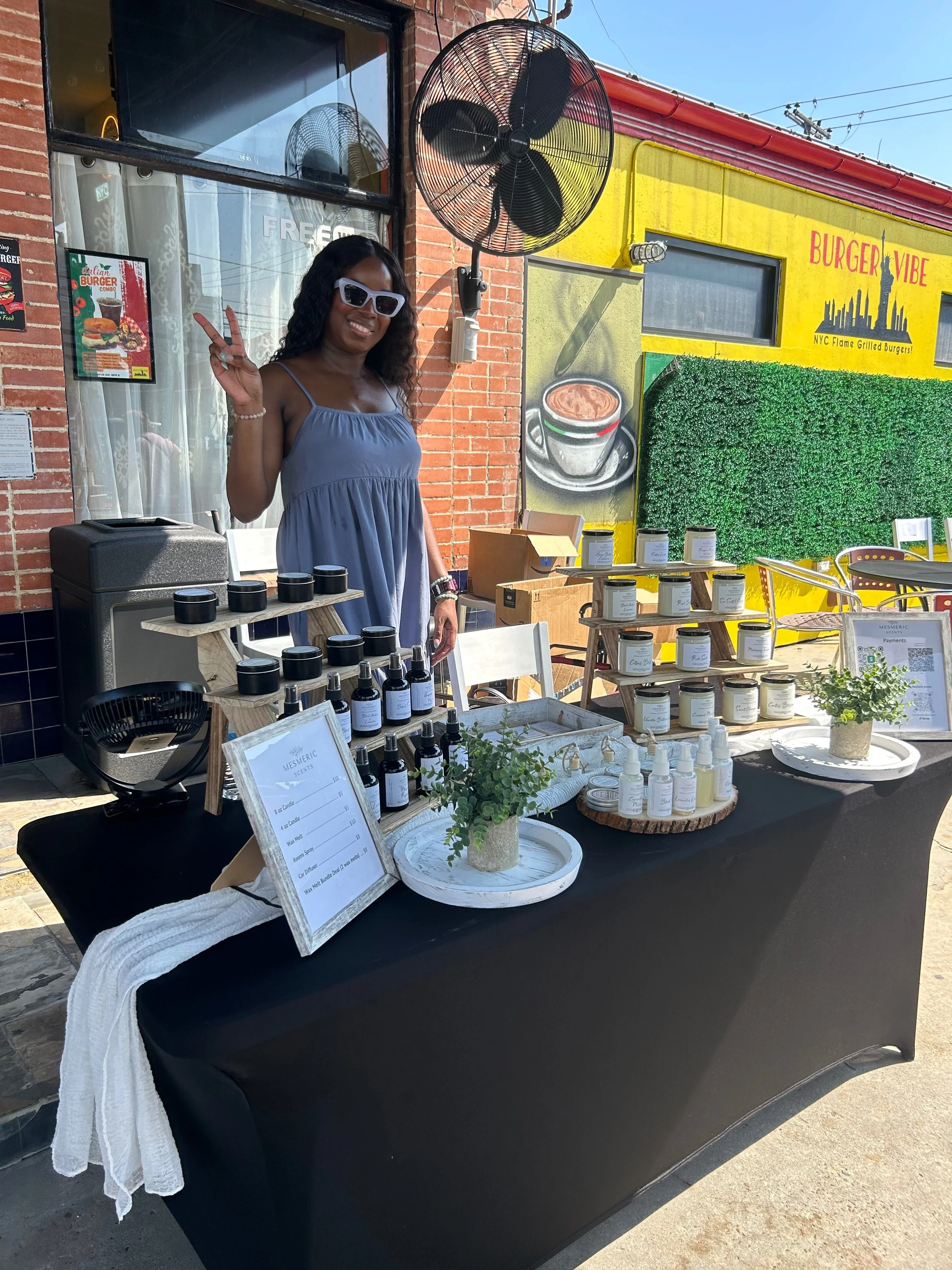 A woman with black curly hair, wearing white sunglasses and a light blue dress, standing behind a table at an outdoor market stall. The table displays various bottles, jars, and plants, with a sign listing prices. The background features a yellow building with a green wall, a coffee cup illustration, and a store sign that reads "BURGER VIBE."