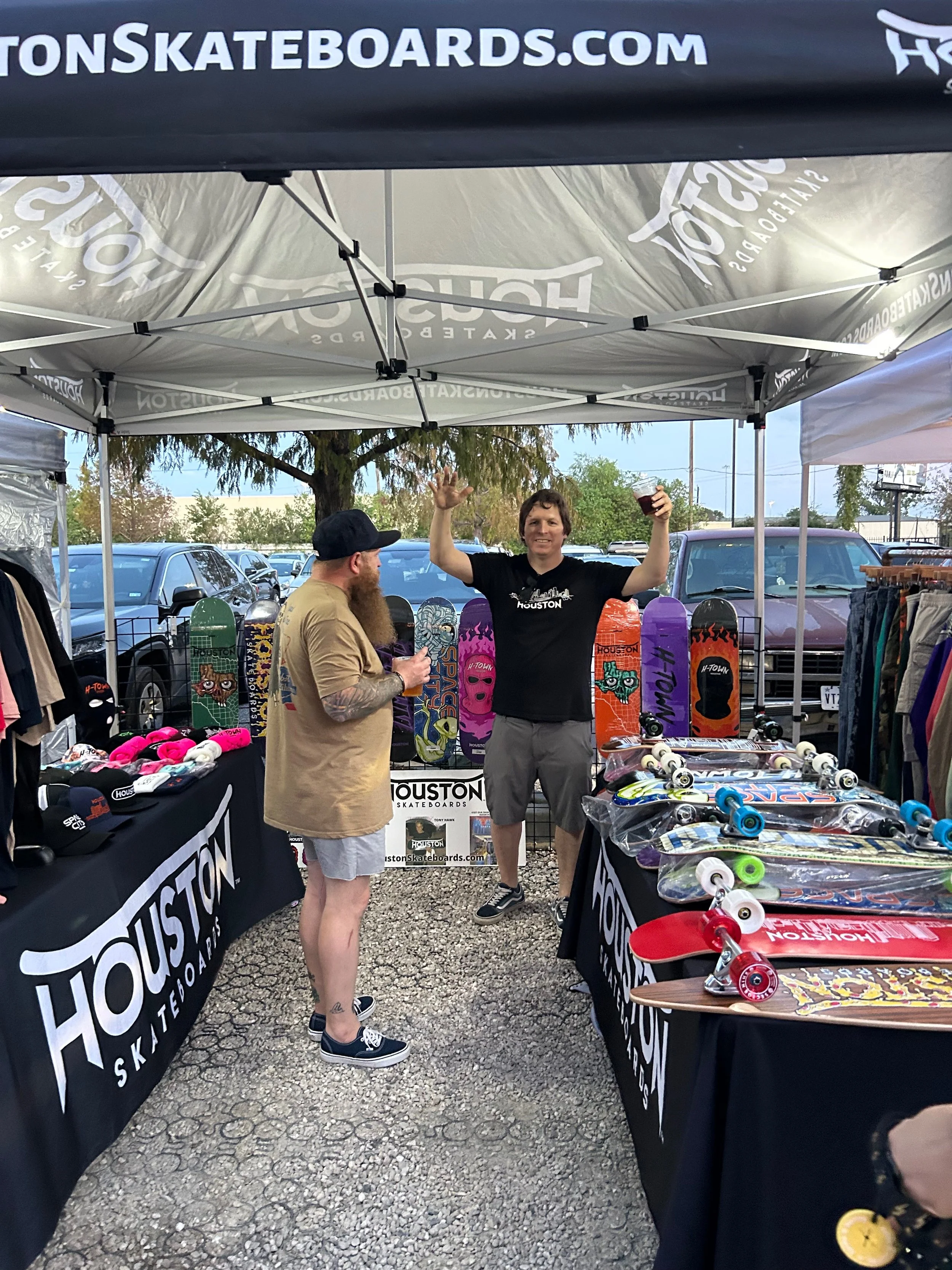 Skateboard booth at an outdoor market with two men, one smiling and holding a drink, surrounded by colorful skateboards, helmets, and skateboarding accessories under a Houston Skateboards tent.