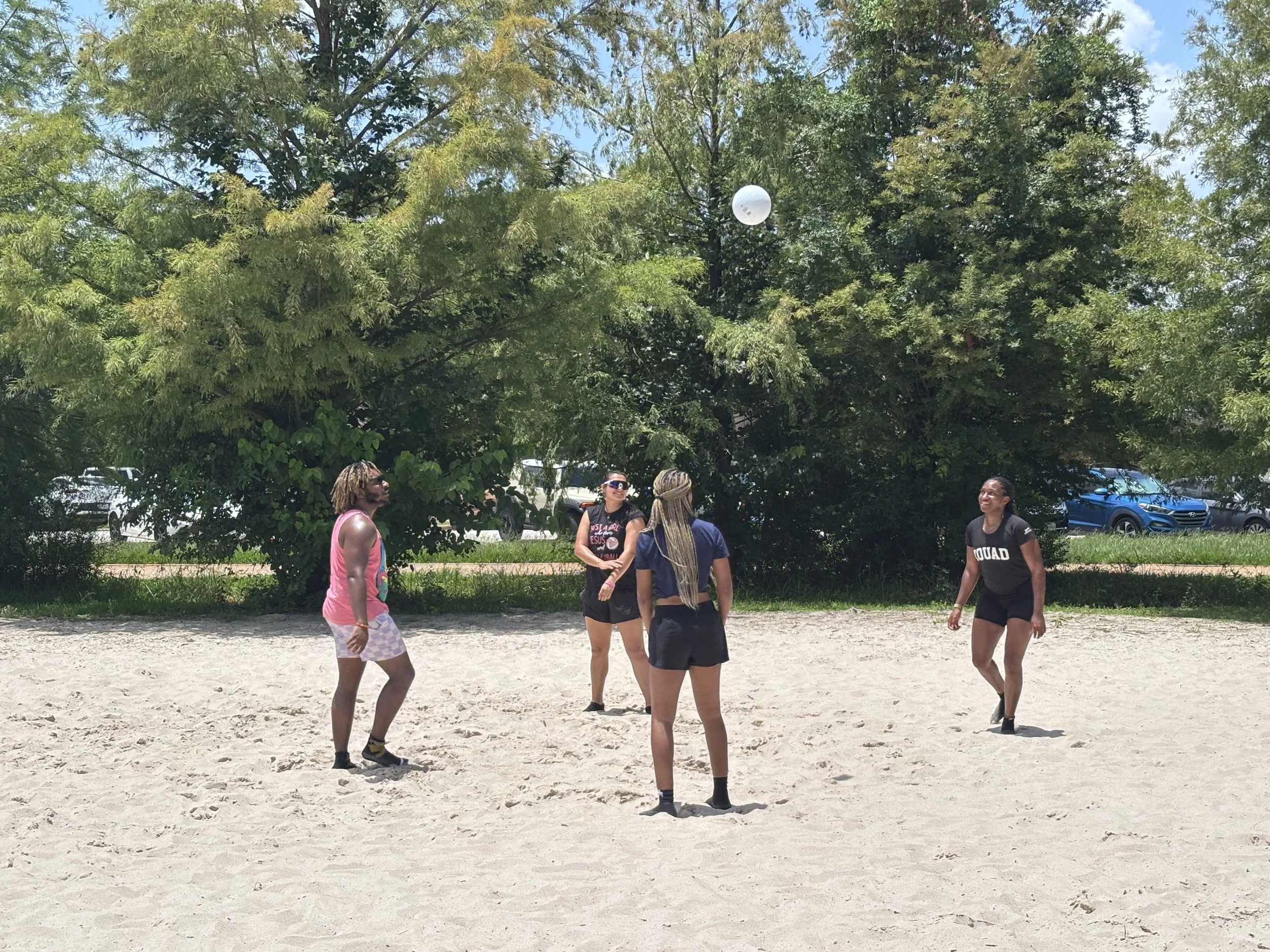 Four women playing volleyball on a sandy beach near trees and parked cars.