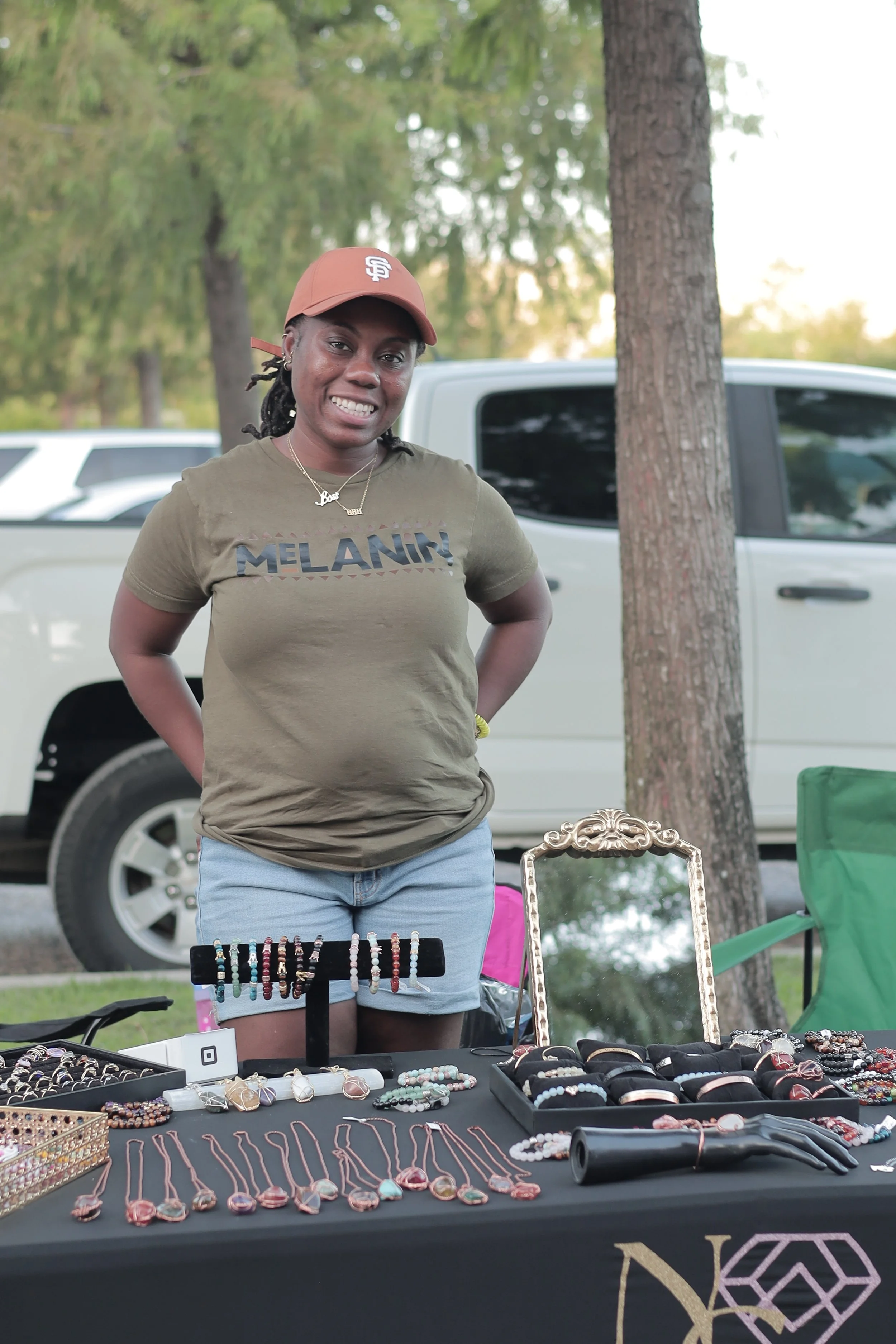 A woman standing behind a table of jewelry at an outdoor market, wearing a tan cap, an olive green T-shirt with "MELANIN" printed on it, and light blue shorts. There are trees, a parked white truck, and some chairs in the background.