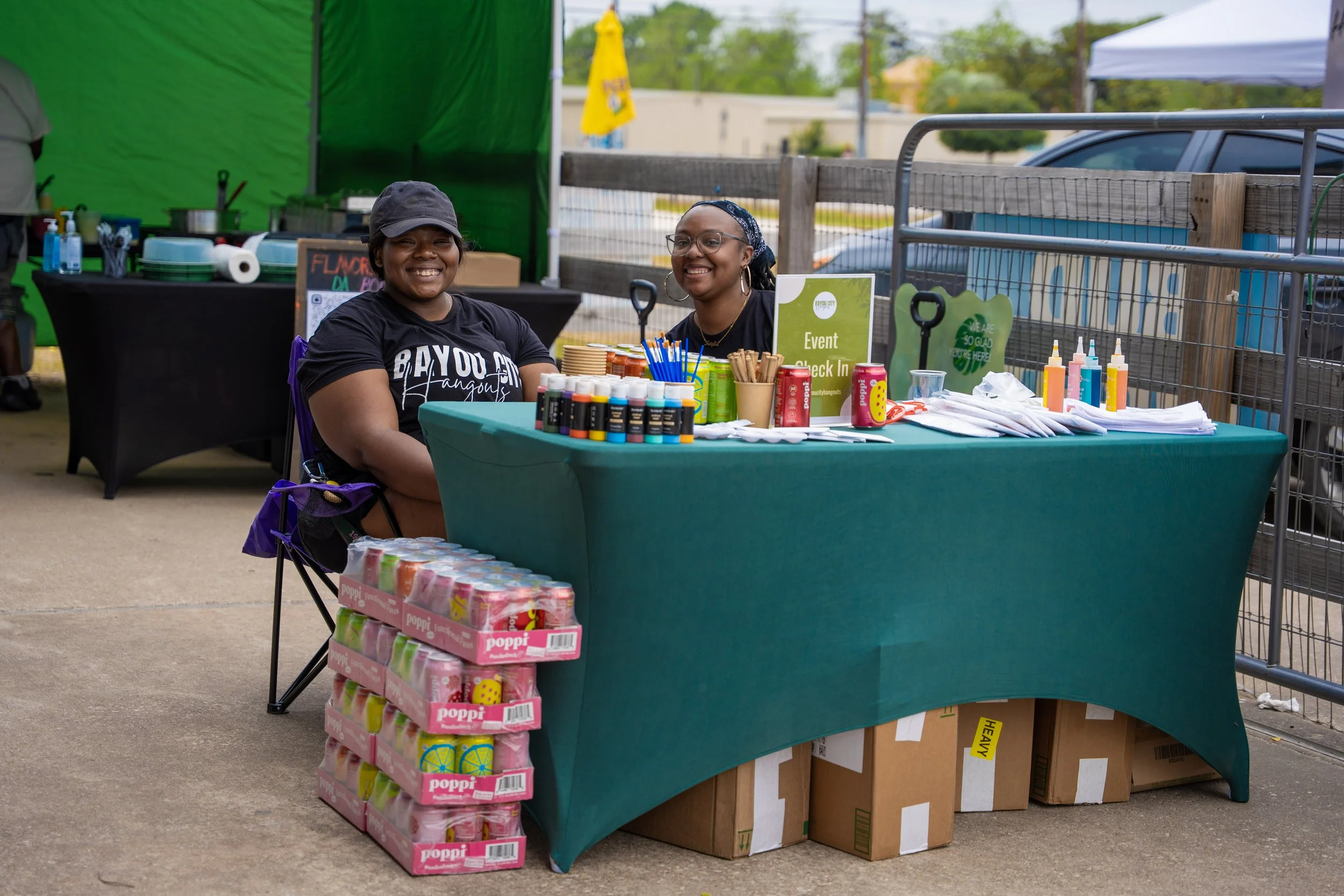 Two women sit at a table with various arts and crafts supplies, including paint bottles, glue, and paper, at an outdoor event. One woman wears a black cap and shirt, smiling, while the other wears glasses and a bandana, also smiling.