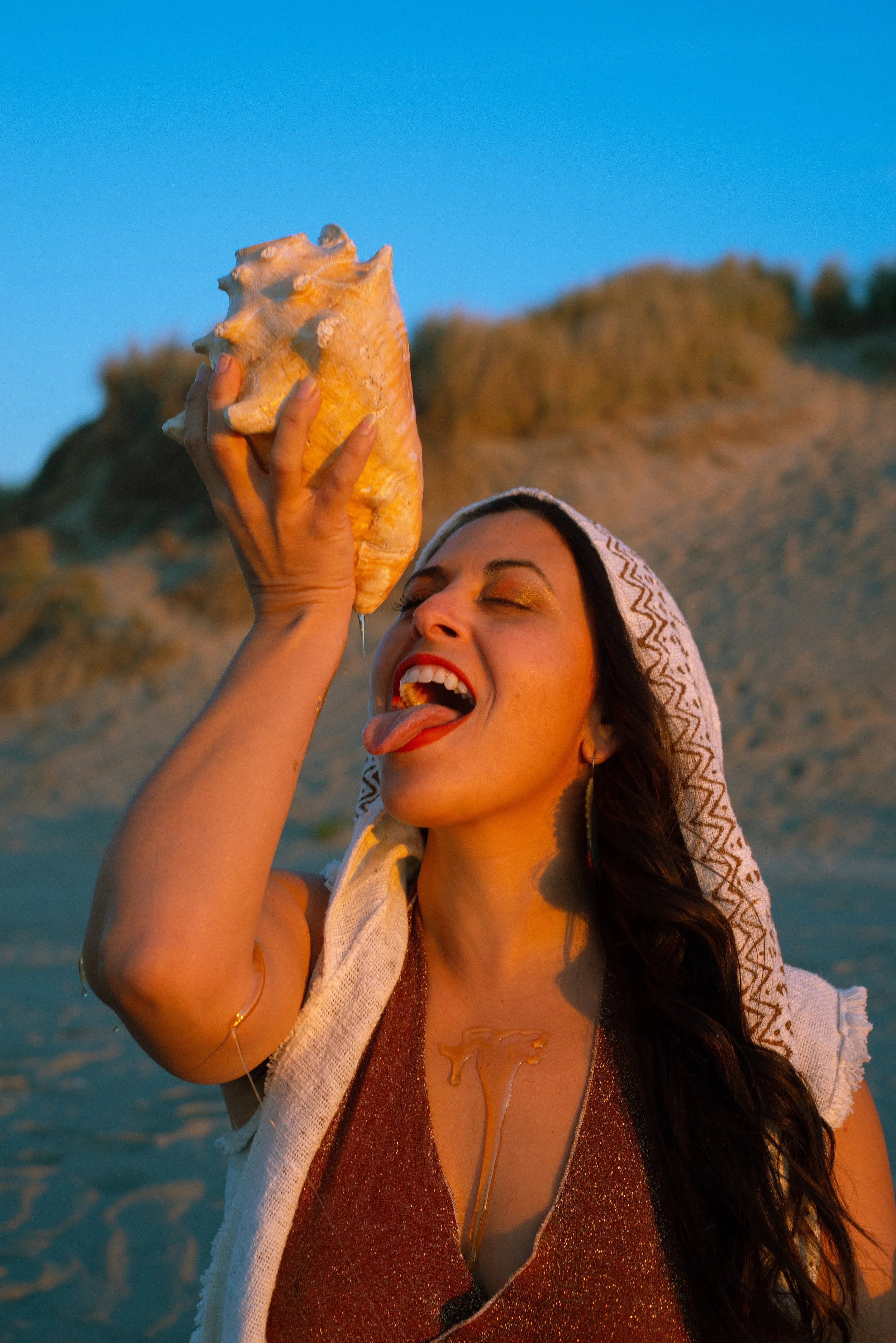 woman dripping honey off of a conch shell, mouth open in anticipation to receive the honey in a playful manner