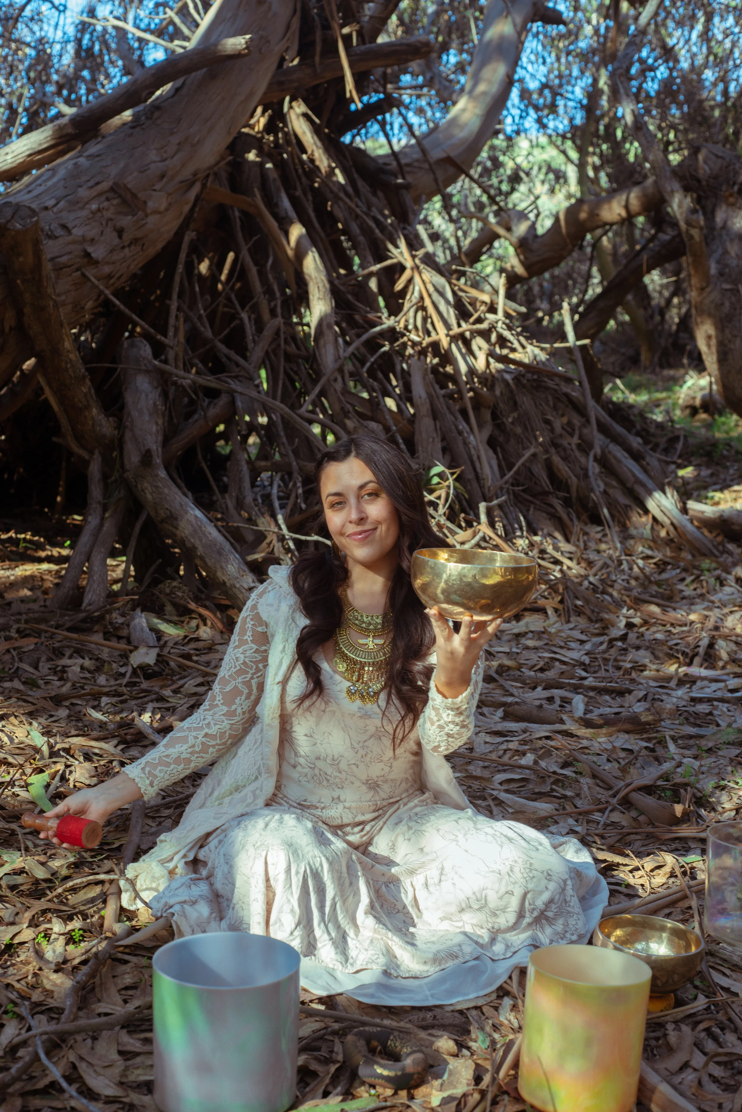 A woman in a white lace dress sitting on the ground among fallen leaves under a tree, holding a golden singing bowl, with other bowls and instruments around her.