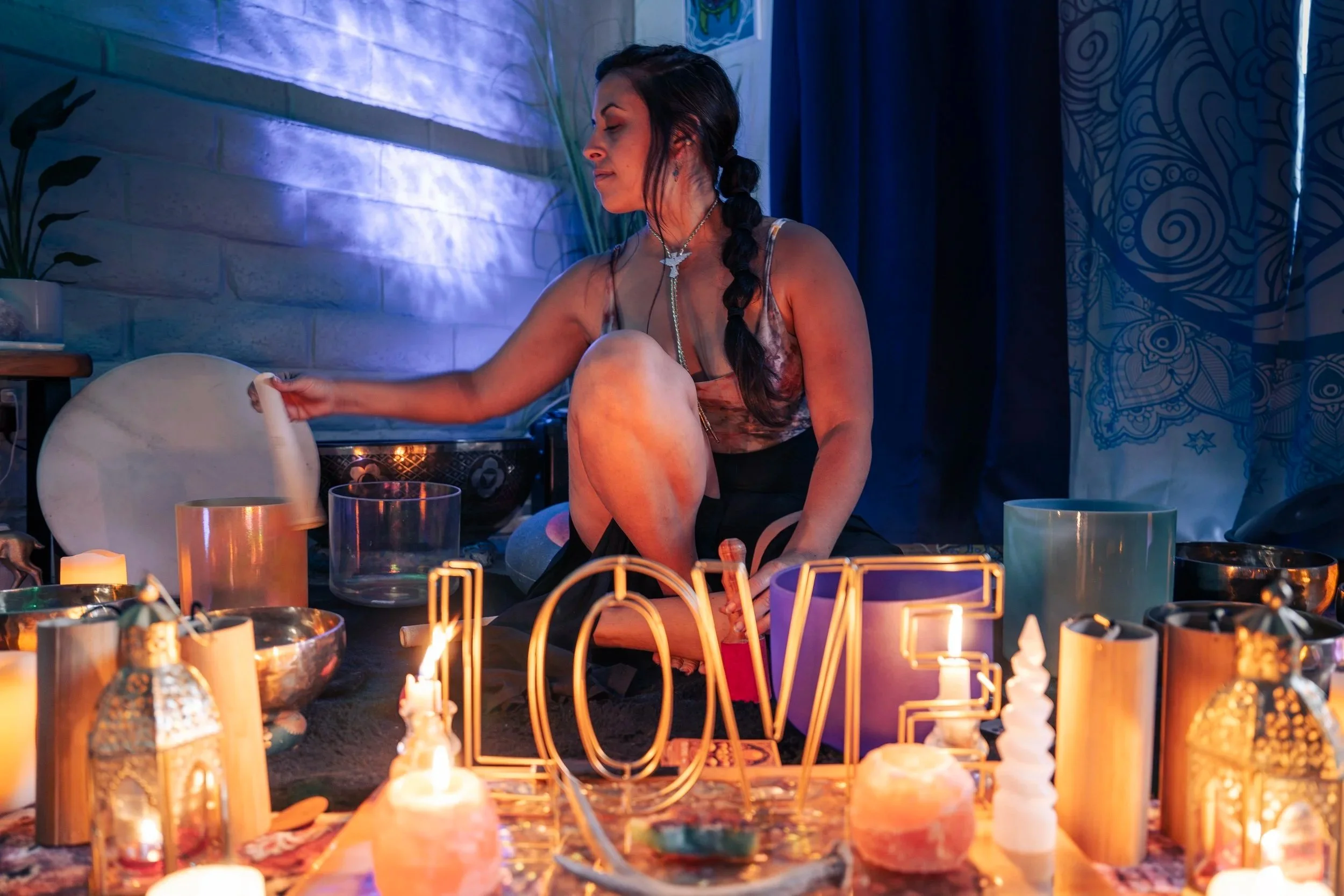 A woman with dark hair in a braid, wearing a sleeveless top, sitting on the floor in a dimly lit room decorated with candles, crystals, and signs that spell 'LOVE' in neon. She appears to be playing a singing bowl surrounded by various decorative objects.