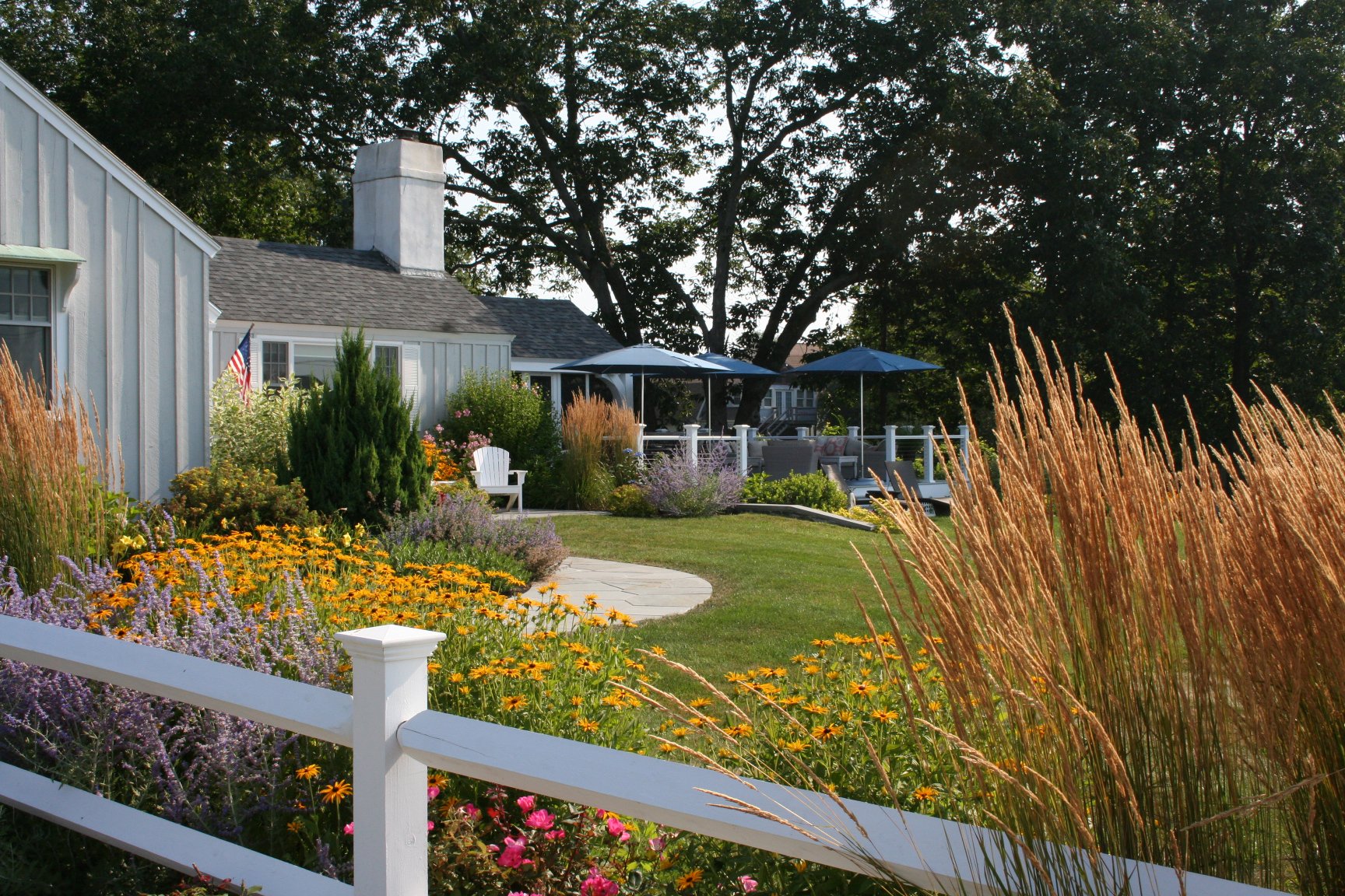 Seabreeze Cottage features a charming white picket fence, lush coastal plantings, and a welcoming New England landscape designed to enhance curb appeal and outdoor living. This timeless residential landscape by Sea Laurel Landscape Architecture blend