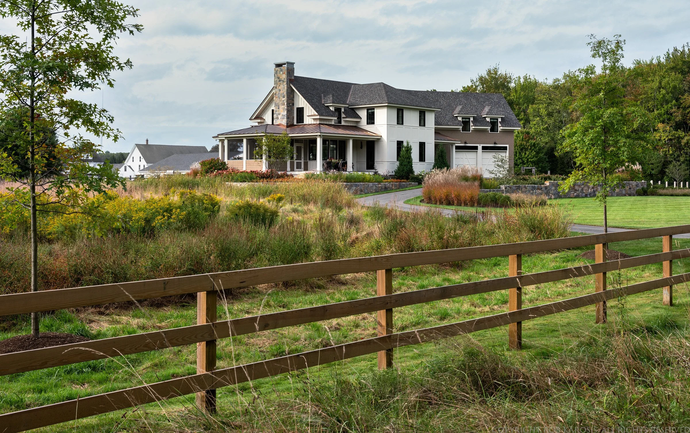A large, modern house with a gray roof and white exterior, surrounded by a landscaped yard with green grass, trees, and bushes, with a curved driveway in front.