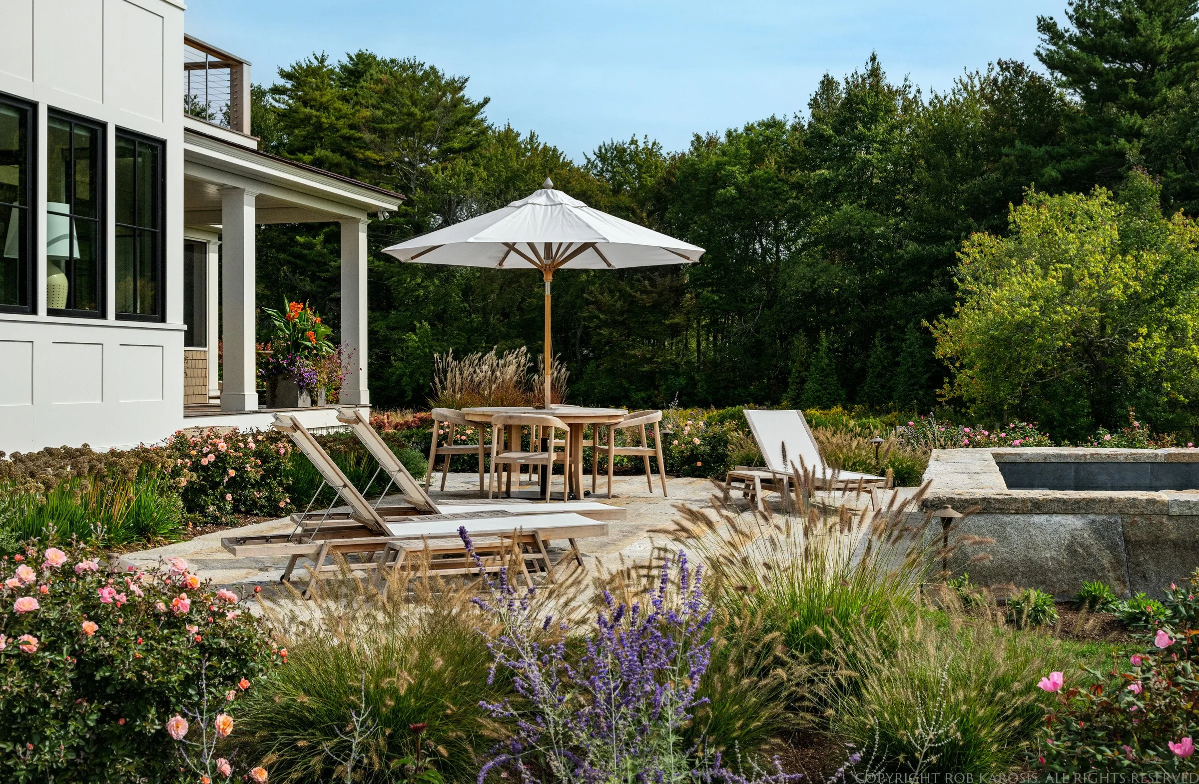 A backyard patio with a white house to the left, outdoor furniture, a large white umbrella, lounge chairs, a flower garden, and a lush green tree background.