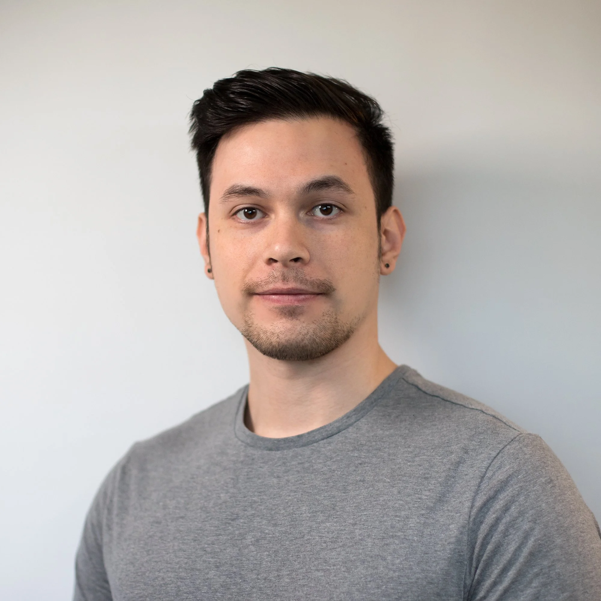 A young man with short dark hair, light facial hair, wearing a gray t-shirt, standing against a plain white background.