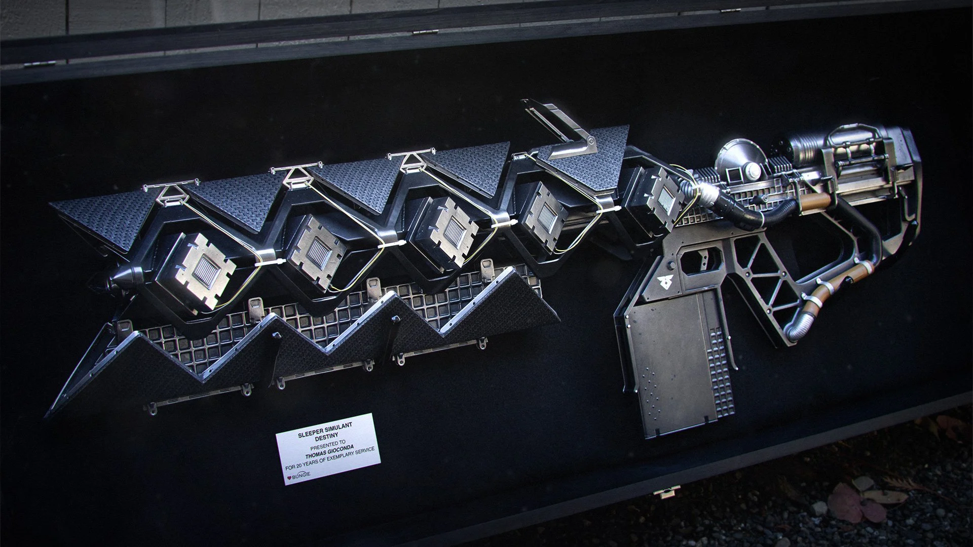 A detailed sci-fi-style weapon or device with multiple segments, pipes, and metallic components, displayed on a black surface with a small informational plaque.