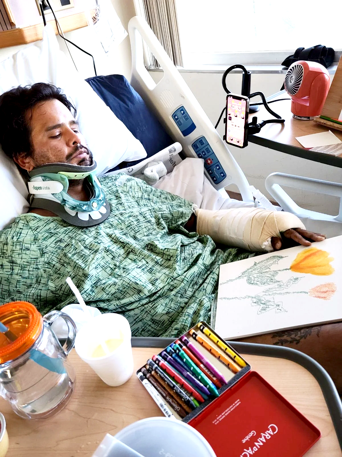 A man in a hospital bed with a neck brace and arm cast, lying with his eyes closed. His room has medical equipment, a bedside table with drawing supplies including colored pencils and paper, and a window with curtains.