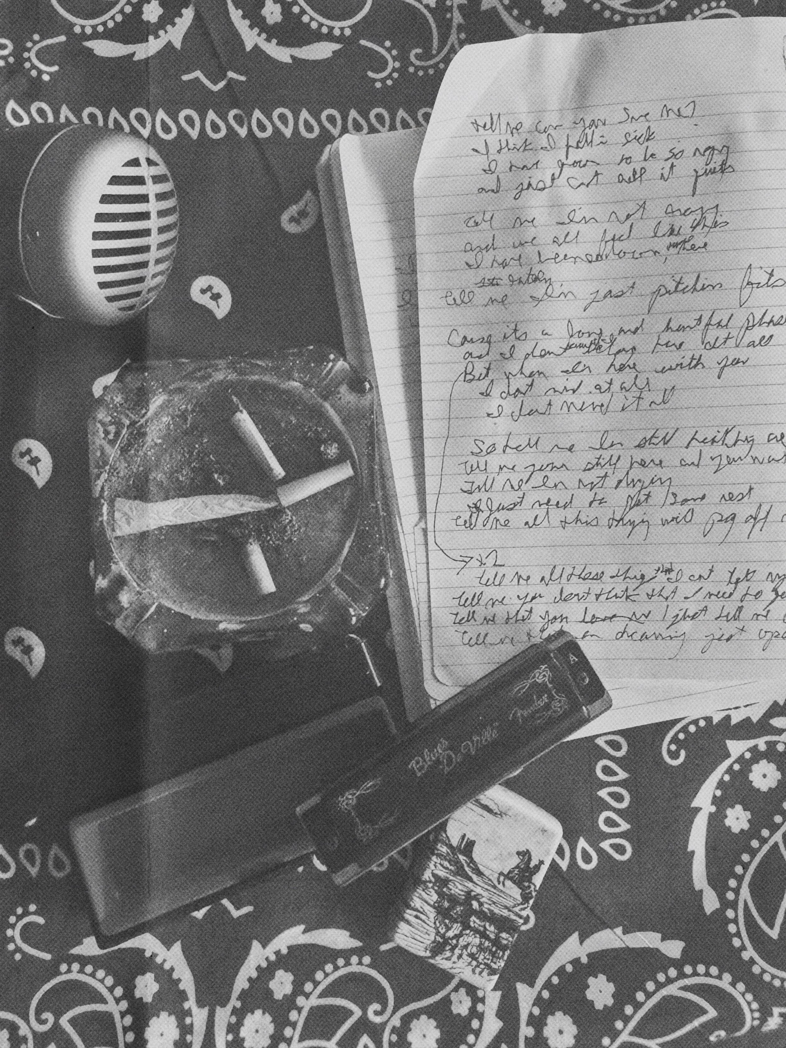 A black-and-white photo of a bandana with a paisley pattern, a pack of cigarettes, an ashtray with cigarettes, a lighter, a handwritten note by Corey La Rue with cursive writing, and a harmonica