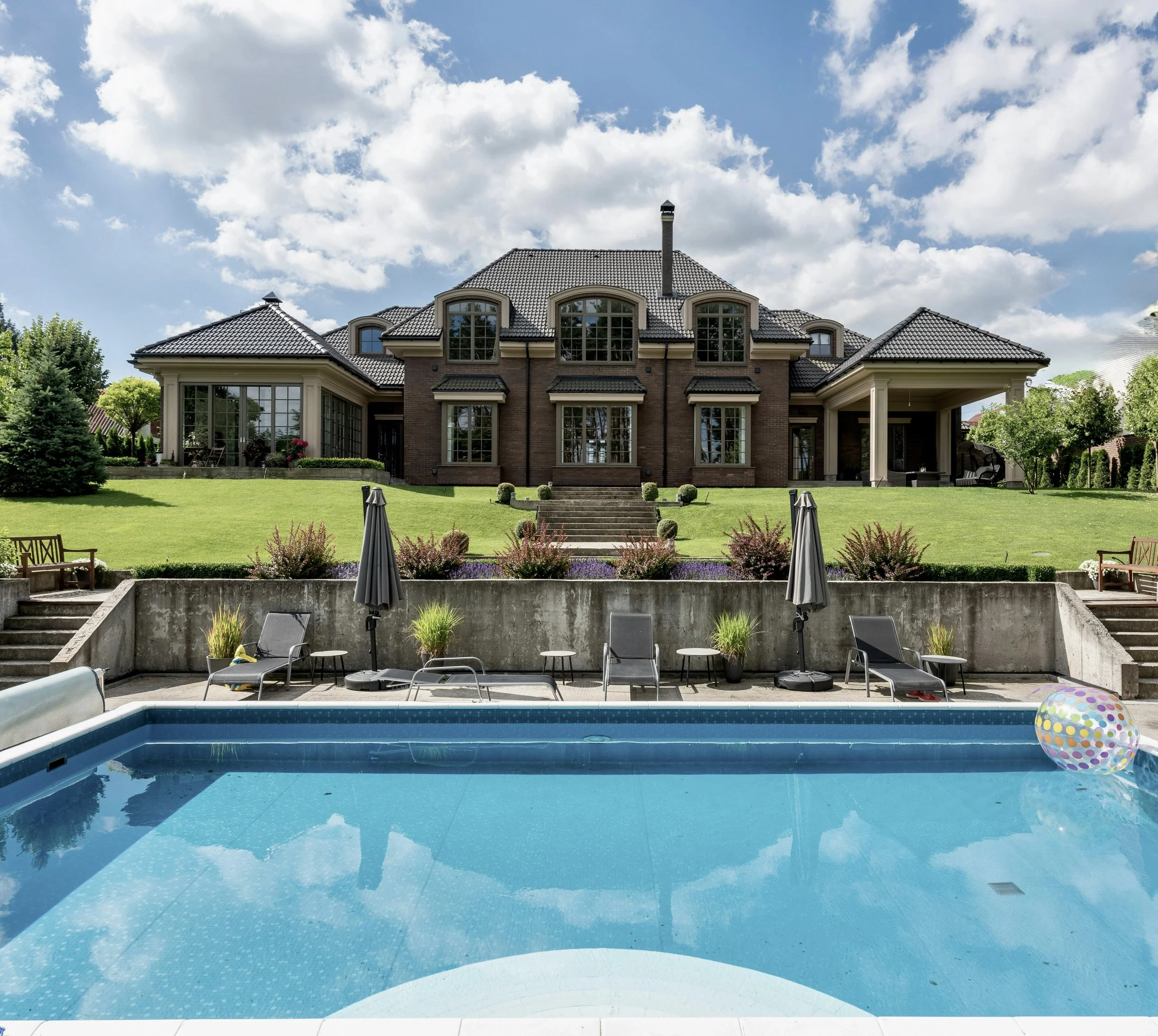 Large house with a brick exterior, multiple windows, and a steep roof, overlooking a well-maintained lawn and a swimming pool with lounge chairs and umbrellas.