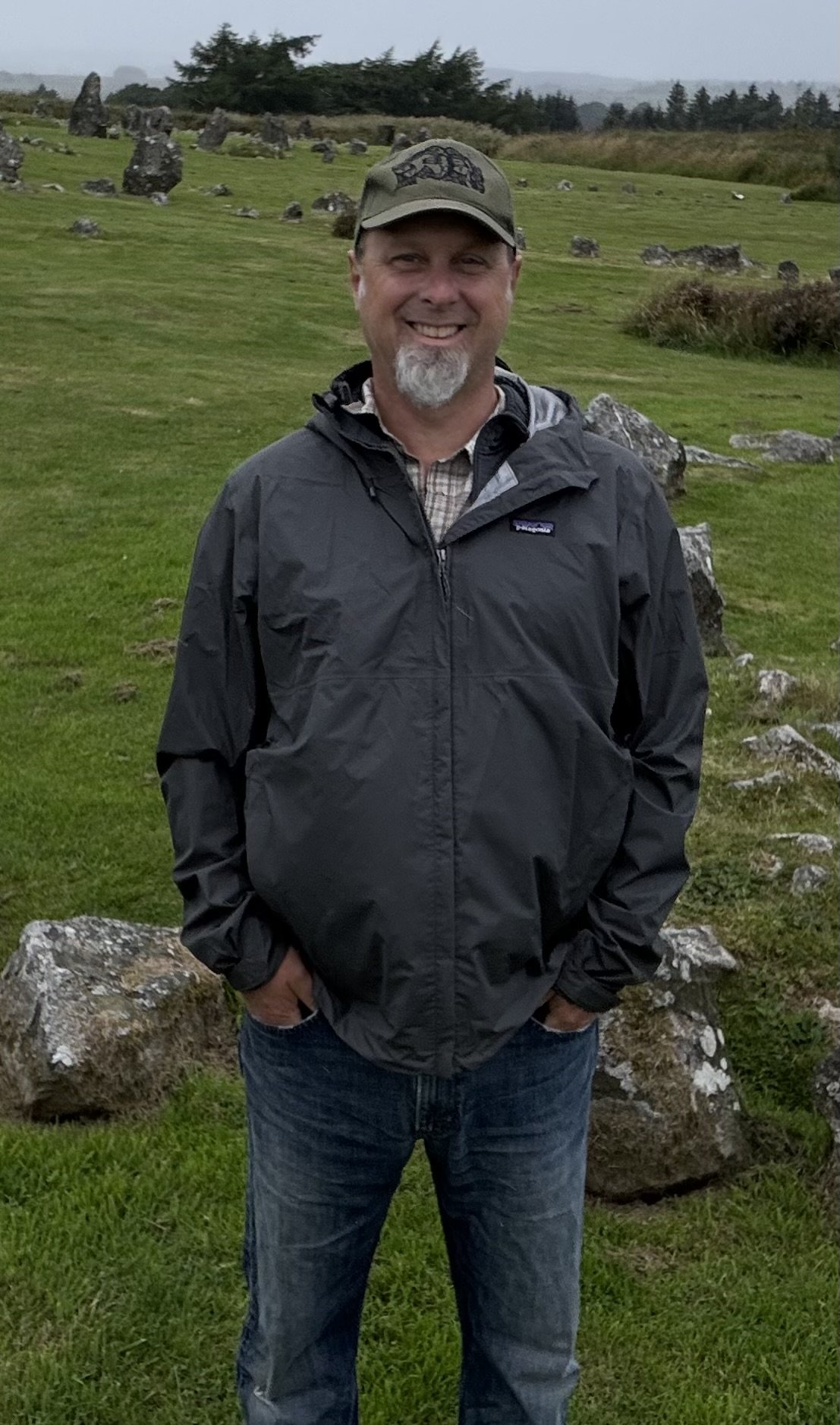 A man standing outdoors on a grassy field with rocks, wearing a gray jacket, blue jeans, and a hat, smiling at the camera.