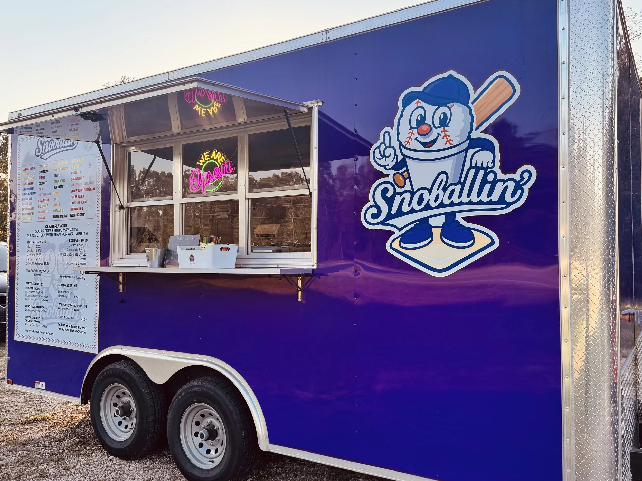 Blue ice cream truck with a cartoon ice cream character holding a baseball bat, wearing shoes and a baseball cap, with the word 'Snoballin'' written below. Neon sign says 'We are Open' and a menu is seen on the side of the truck.