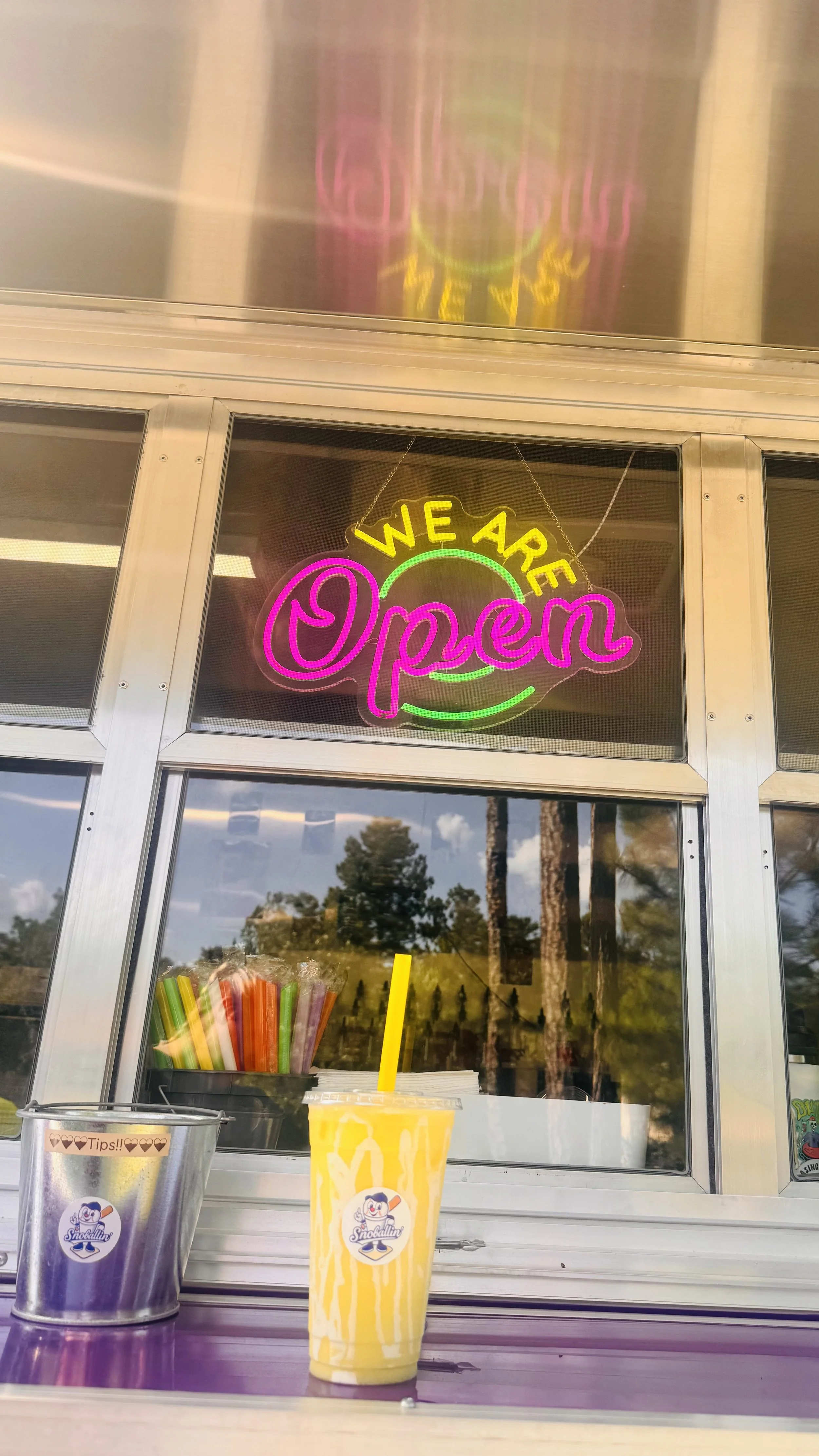 A neon sign reads 'We Are Open' in pink, yellow, and green. A yellow and white beverage with a straw and a logo is placed on a counter in front of a window, with trees and sky visible outside.