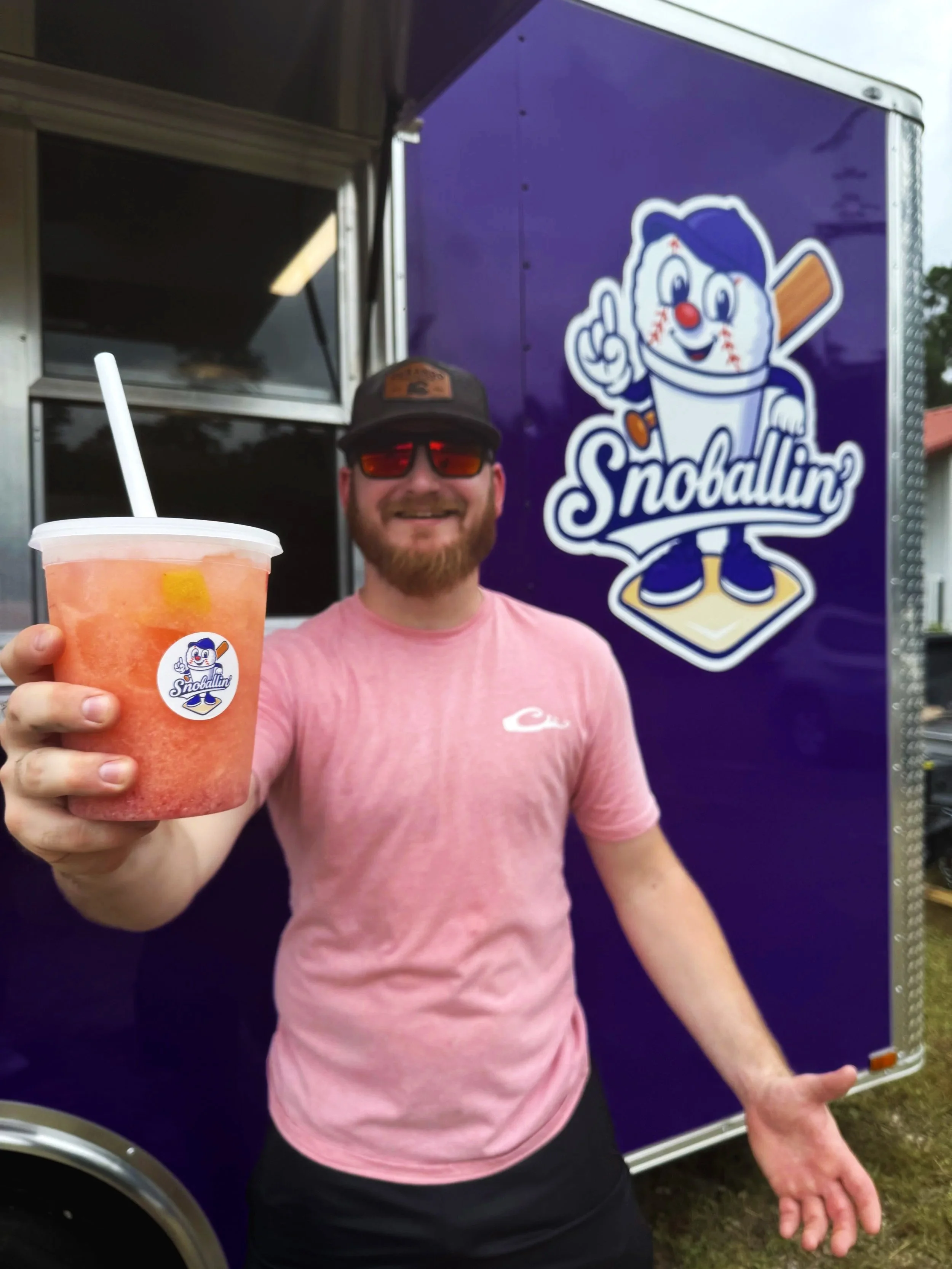Man holding a pink fruit slushie drink with a straw in front of a purple food truck with the logo 'Snoballin' featuring a cartoon snow cone character.