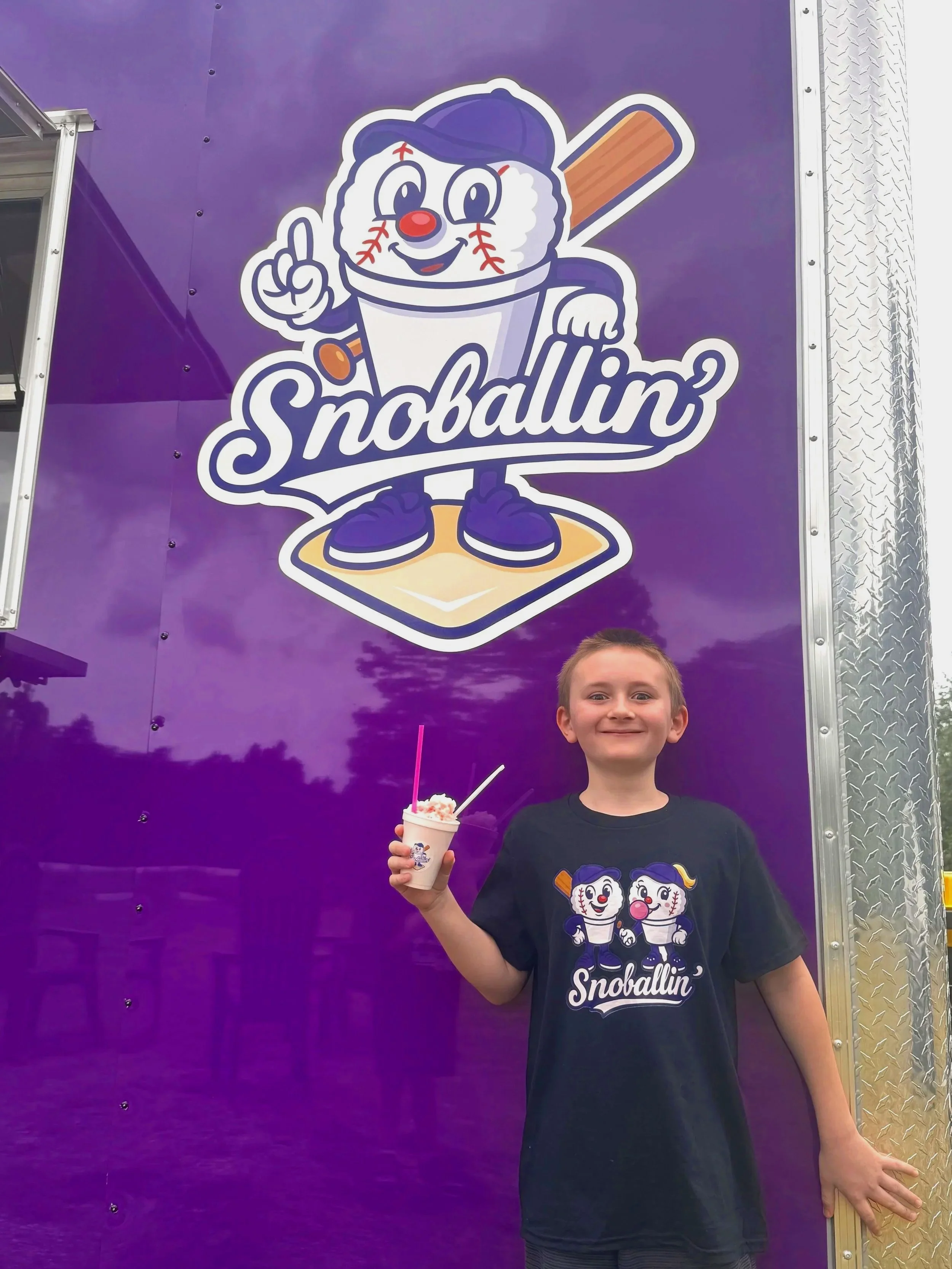 A young boy holding a cup of ice cream, standing in front of a purple board with a cartoon mascot and the word 'Snoballin' on it, wearing a black t-shirt with the same mascot. The mascot is a baseball with a face, wearing a baseball cap and sneakers,